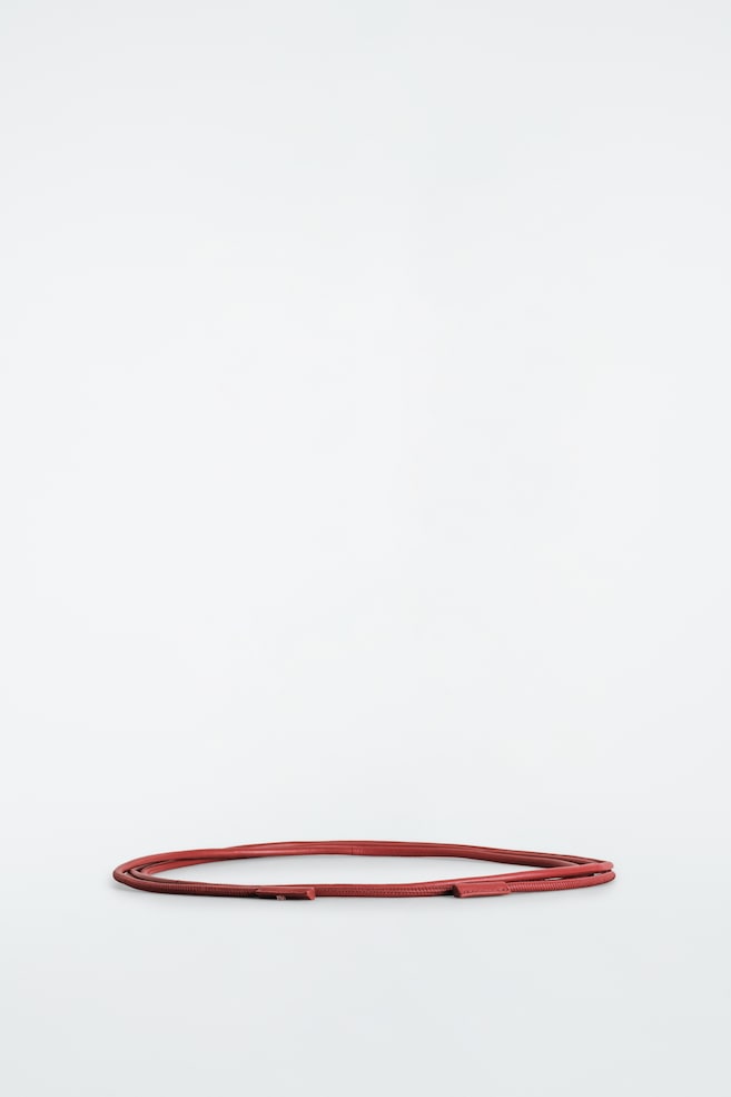 De leren touwriem ligt opgerold in een platte horizontale lus en laat het slanke, afgeronde ontwerp in een roodbruine kleur zien. Fijne stikdetails lopen over de hele lengte, met kleine bijpassende leren eindstukken die de uiteinden afwerken. - 1