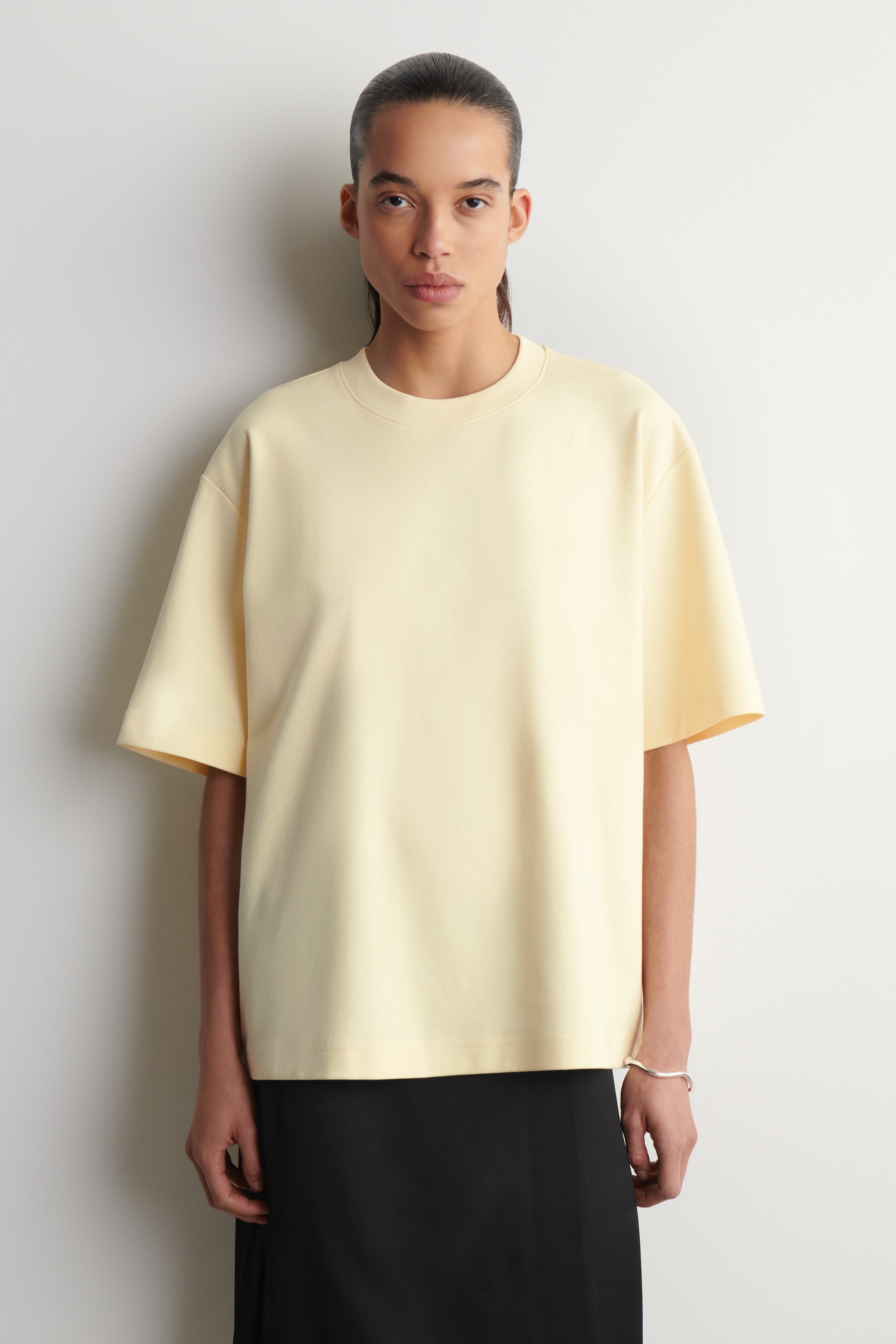 Visualizza immagine più grande: Una t-shirt giallo pallido, dal taglio ampio, viene presentata, caratterizzata da un girocollo, maniche corte larghe che scendono fino al gomito e un orlo dritto, indossata sopra una gonna nera.