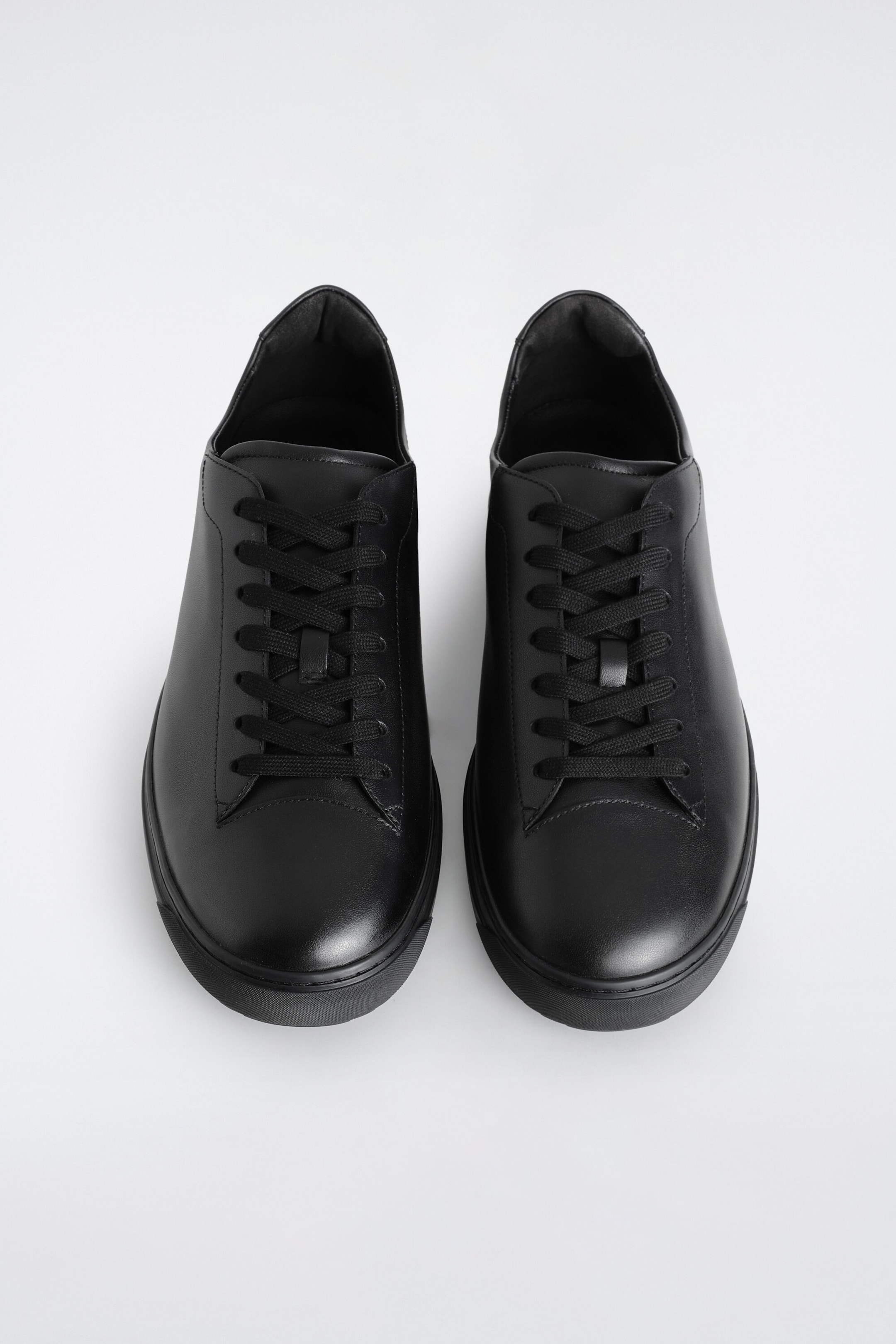 Leather sneakers are shown in a top-down front view, highlighting a minimalist low-top design in smooth black leather. The shoes feature matching black laces, rounded toes, and thick black rubber soles. Leather sneakers are shown in a top-down front view, highlighting a minimalist low-top design in smooth black leather. The shoes feature matching black laces, rounded toes, and thick black rubber soles.