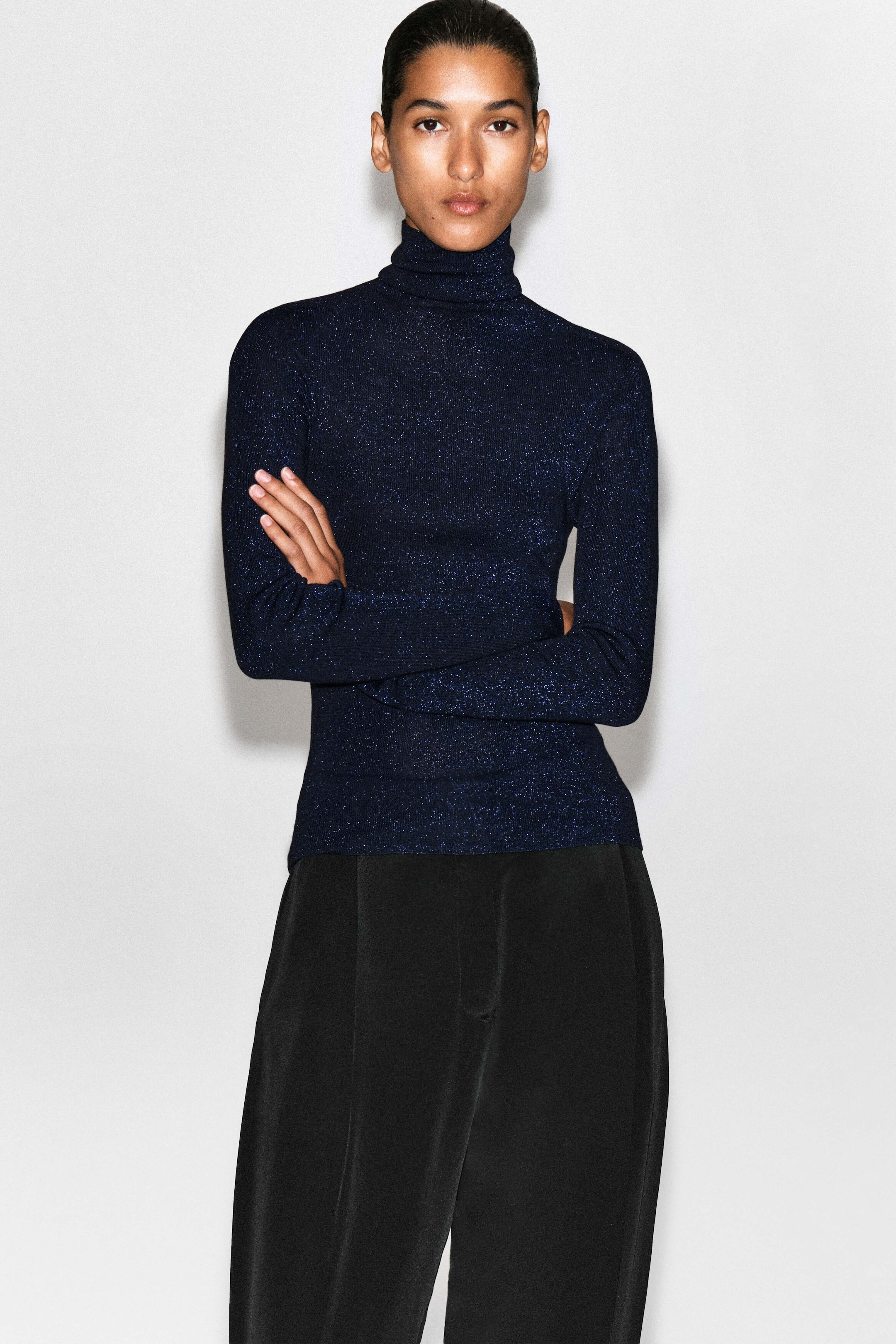The sparkly seamless wool funnel-neck top showcases a slim fit in a dark blue, glittery metallic knit. Facing forward, the long-sleeved top features a high folded collar and rests over black trousers.