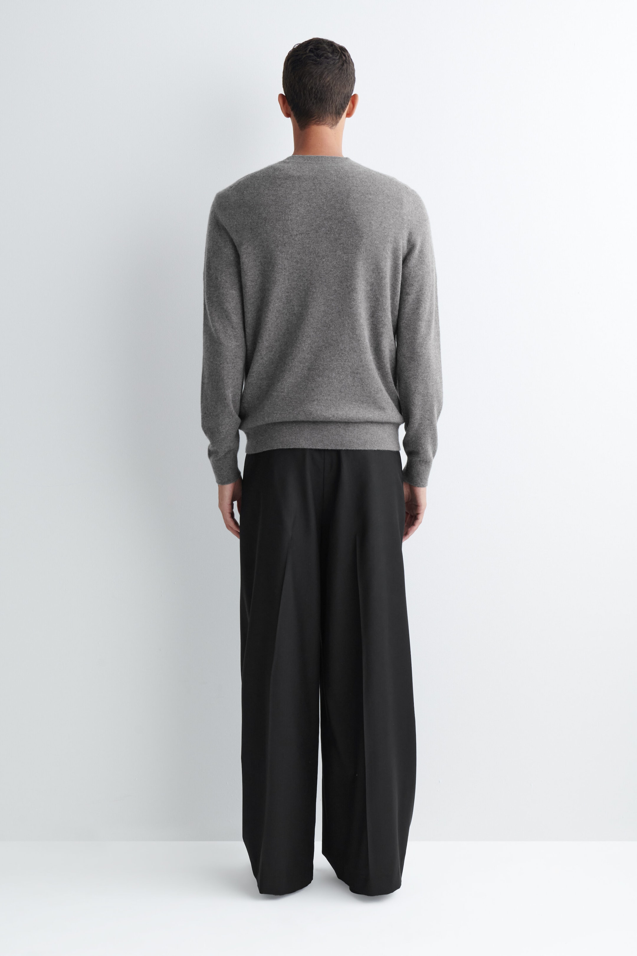 Schwarze, plisseegefaltete Wide-Leg-Hose in lockerer Passform wird vom Model getragen und zeigt eine weite Silhouette mit markanten vertikalen Bügelfalten entlang der Rückseite jedes Hosenbeins. Die Hose ist mit einem langärmeligen grauen Rundhalspullover kombiniert.
