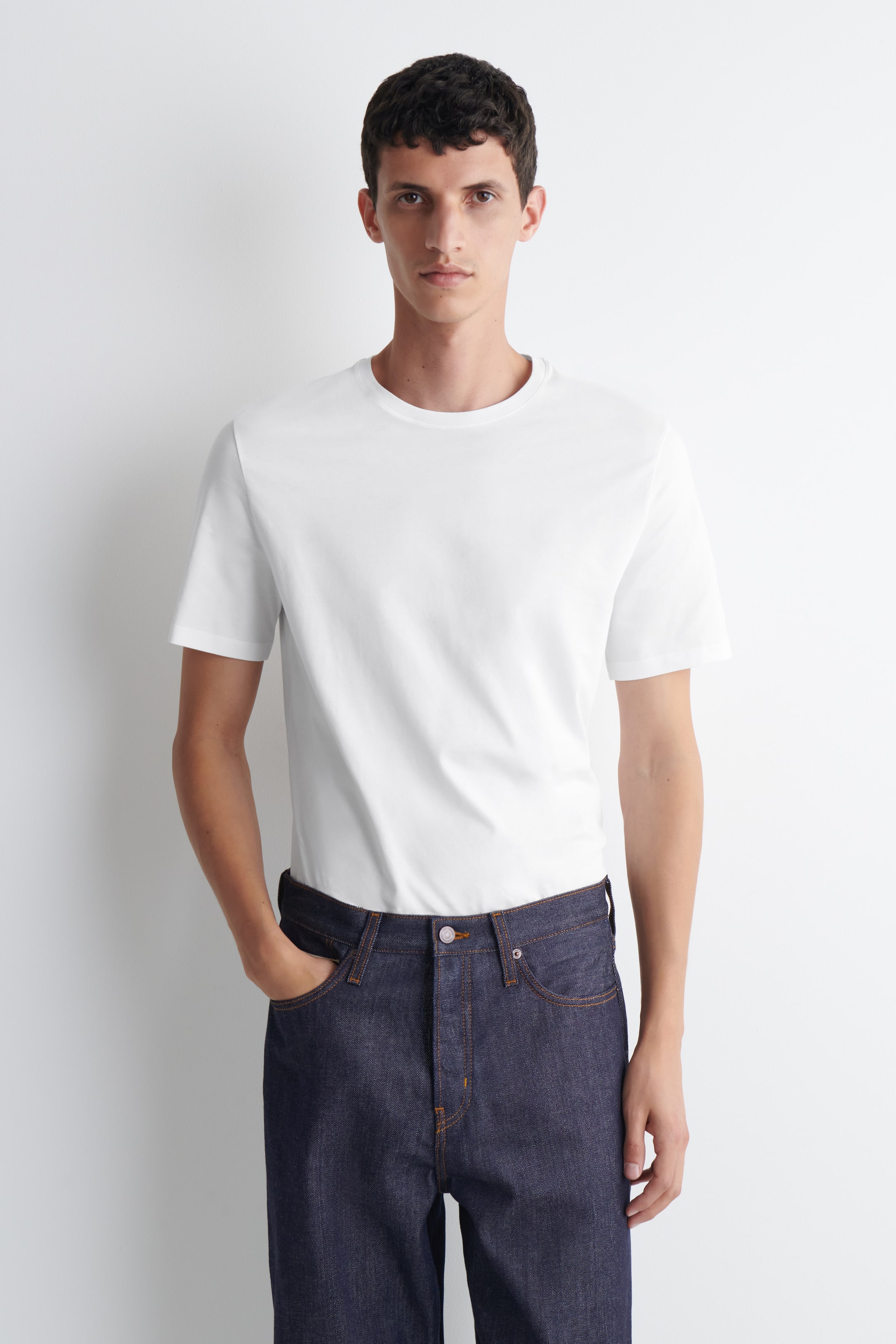 Visa större bild: En vit t-shirt med en klassisk rund hals och korta ärmar, som sitter smidigt över överkroppen och bärs instoppad i mörka jeans.