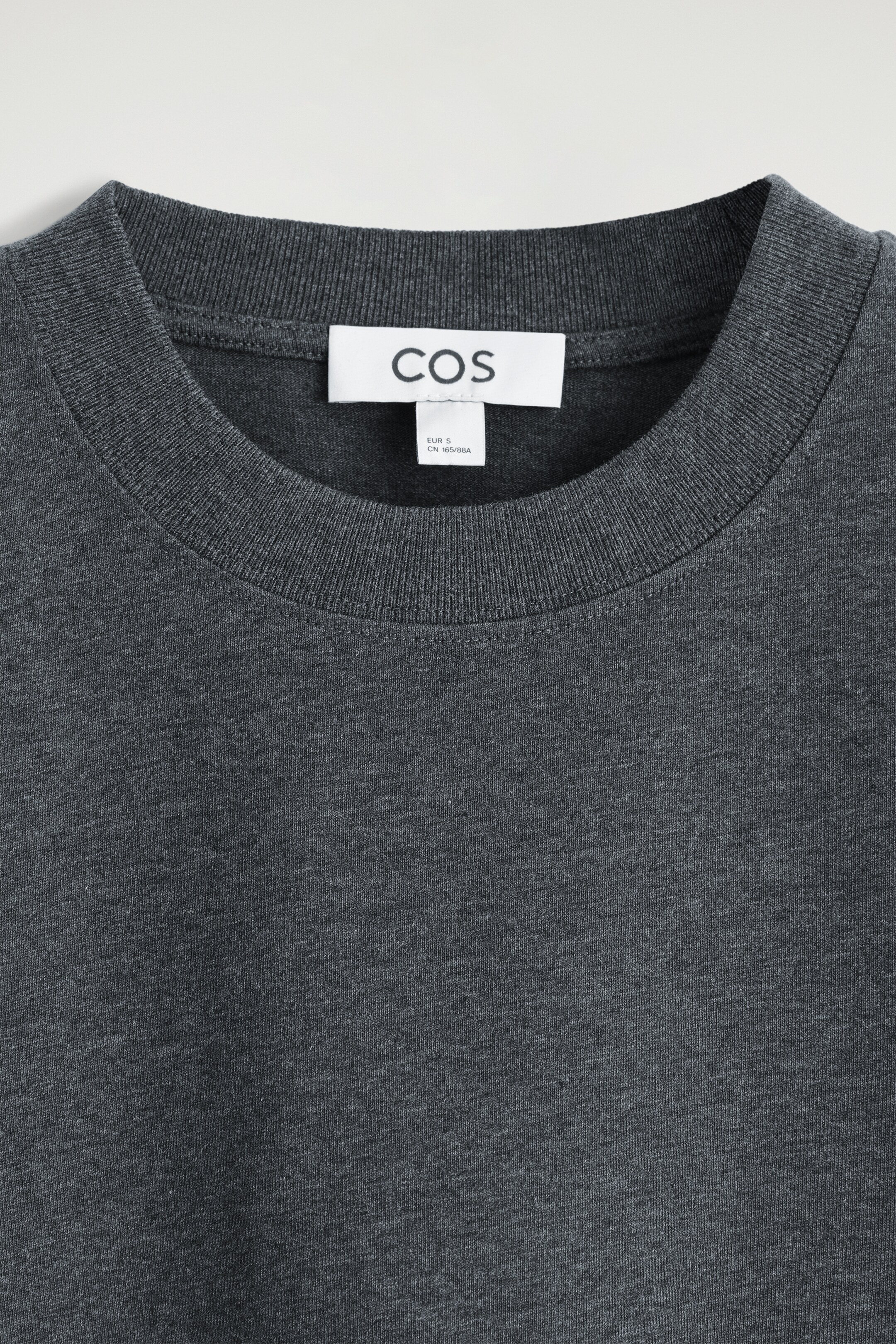 Größeres Bild anzeigen: Ein dunkelgraues T-Shirt wird von vorne gezeigt und präsentiert seinen gerippten Rundhalsausschnitt und den doppelt genähten Halsausschnitt. Ein weißes COS-Markenetikett ist am hinteren Teil des Halses sichtbar, und der Stoff ist ein melierter Jersey.