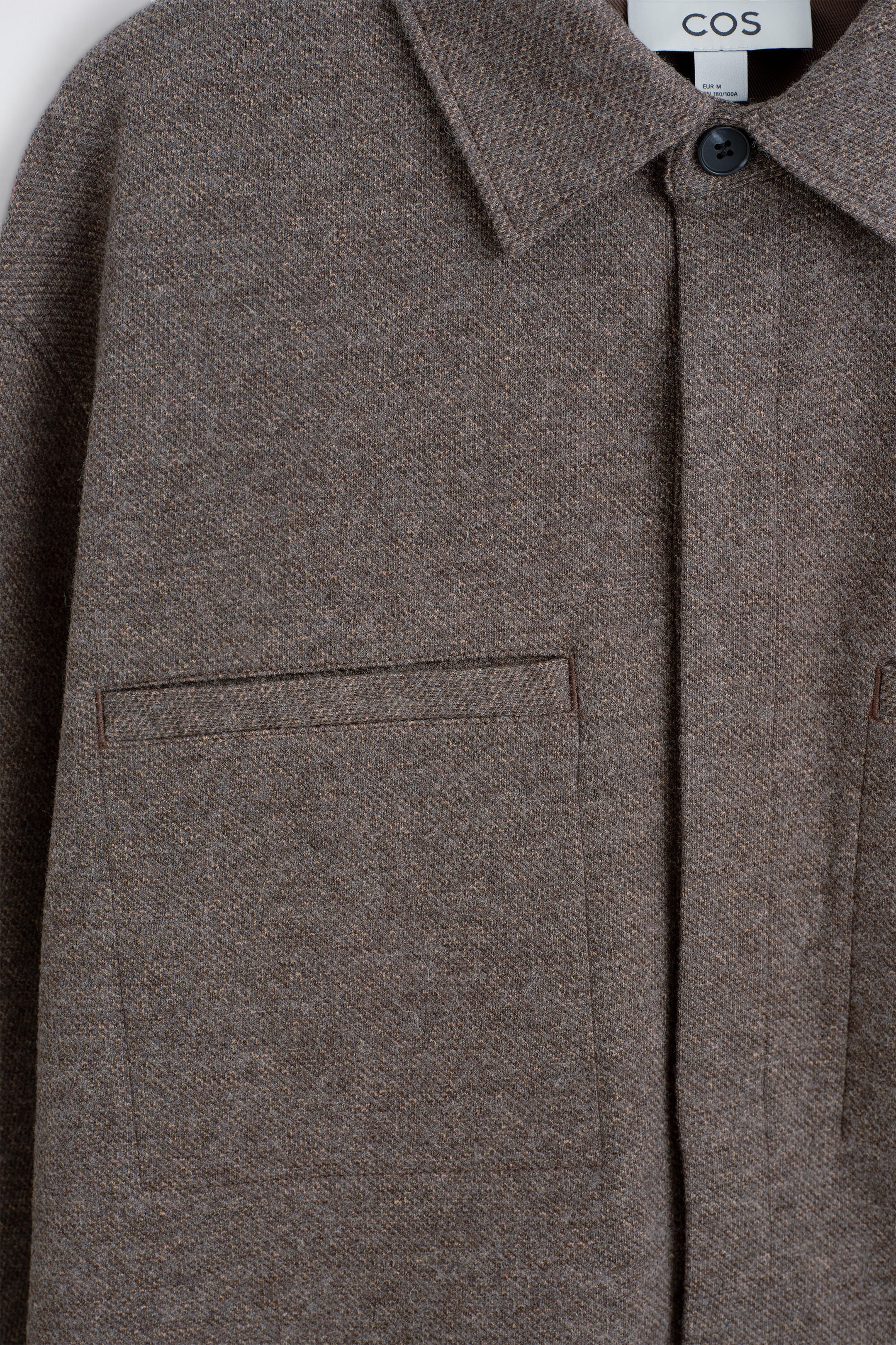 A heathered brown overshirt features a pointed collar with a dark button closure at the top. A white 'COS' brand tag is visible at the neck, and a rectangular flap patch pocket is visible on the left chest. A heathered brown overshirt features a pointed collar with a dark button closure at the top. A white 'COS' brand tag is visible at the neck, and a rectangular flap patch pocket is visible on the left chest.