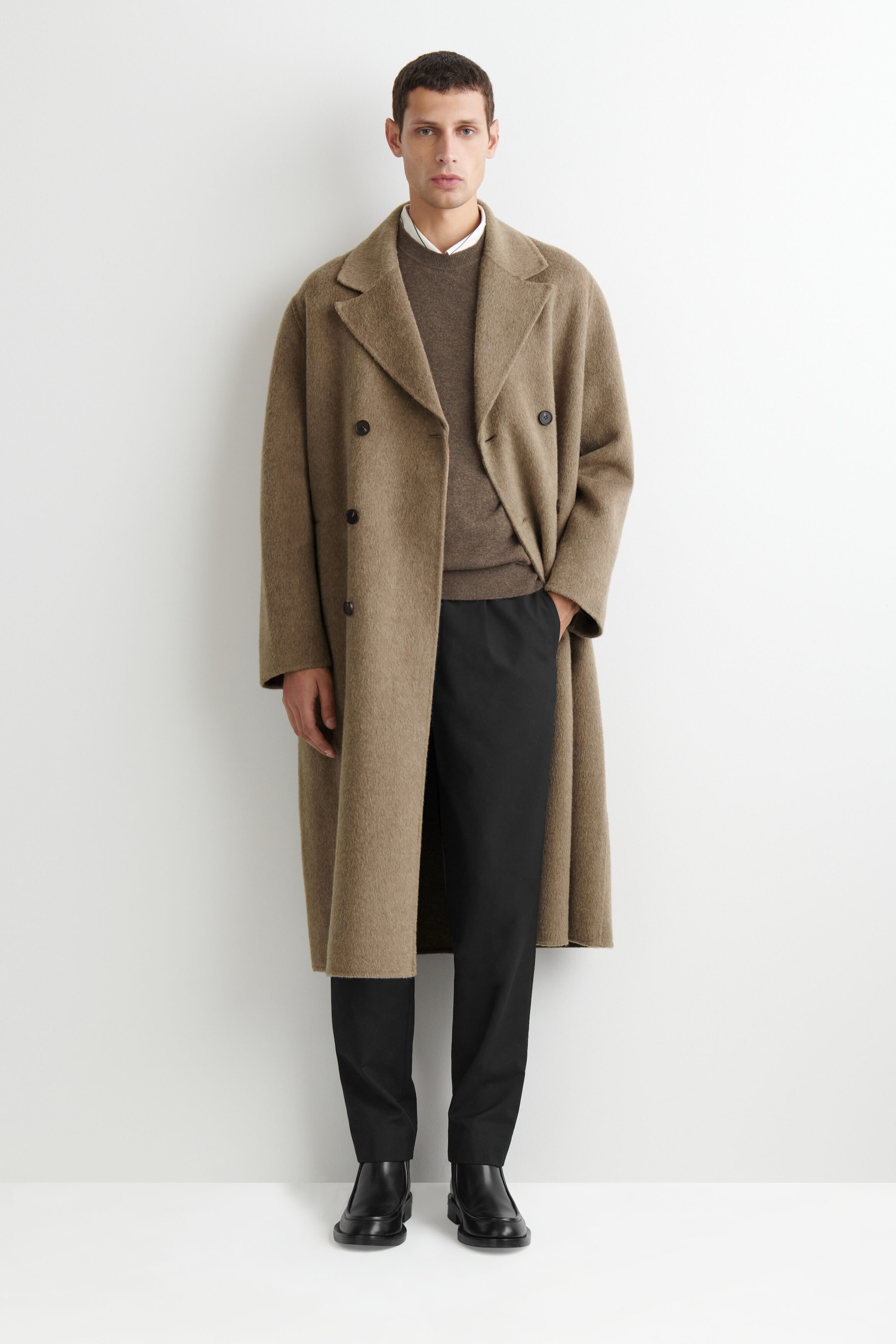The double-faced wool-blend long coat in a taupe-brown hue is shown from the front, featuring wide notch lapels, a double-breasted button design, and a relaxed, straight-cut silhouette. The coat is worn open over a layered brown crewneck sweater, white collared shirt, and black trousers.