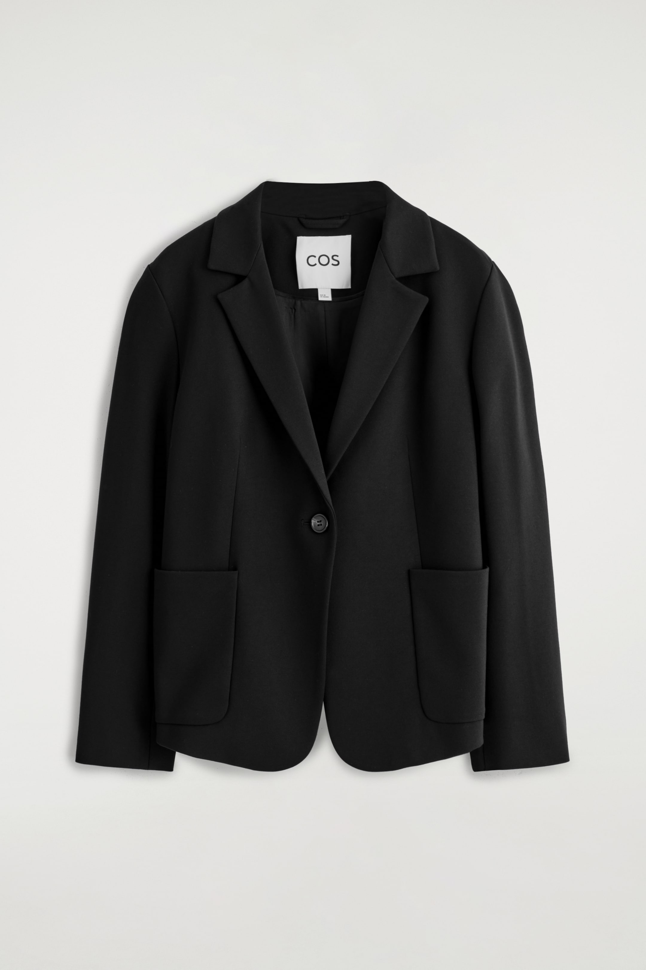 Agrandir l'image: Un blazer noir est présenté de face, avec des revers crantés, une fermeture à un bouton et deux grandes poches plaquées avec des bas arrondis. Il a une texture lisse, un ourlet avant subtilement arrondi, et une étiquette de marque blanche "COS" est visible à l'arrière du cou.