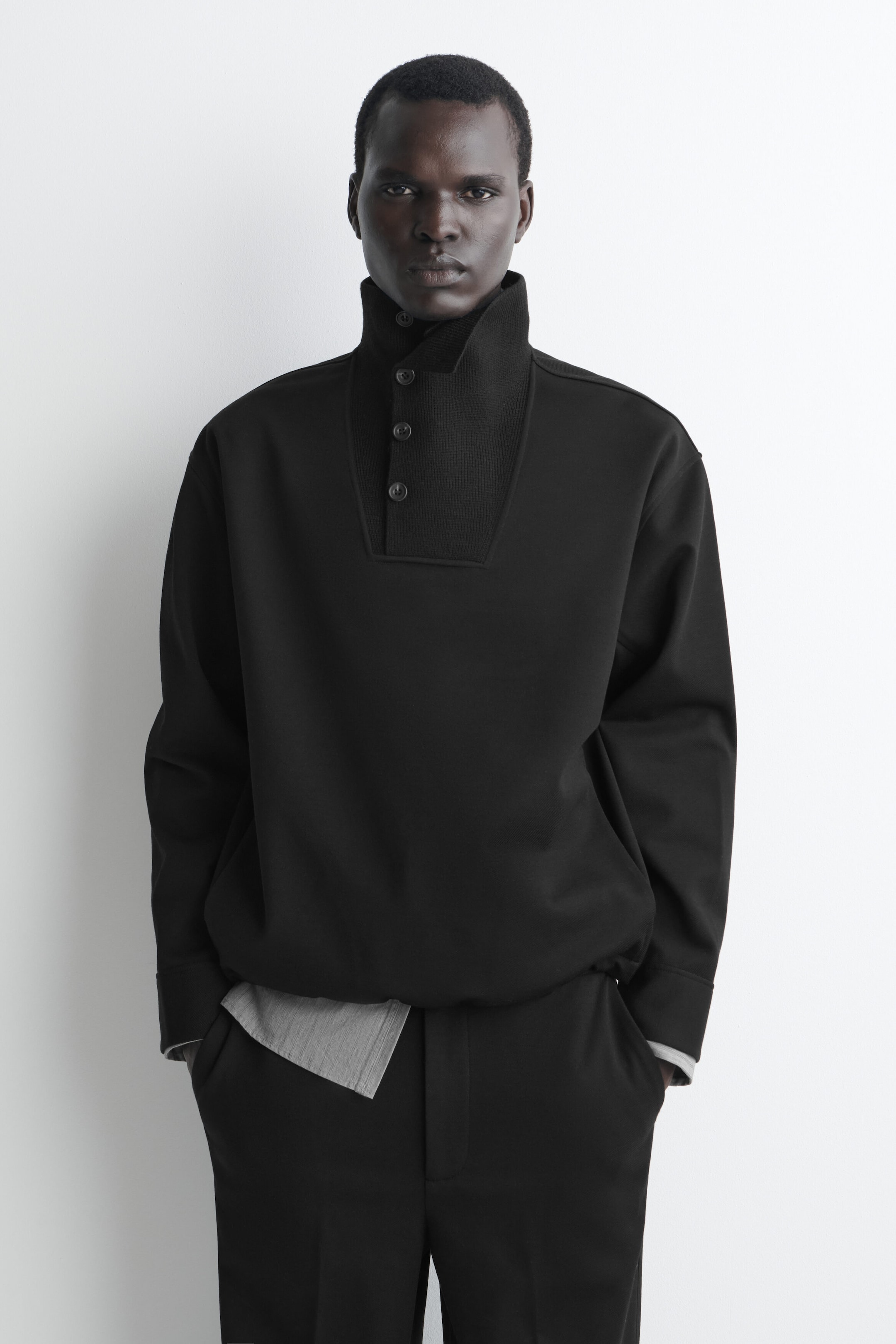 A black high-neck sweatshirt is shown from the front, featuring a relaxed fit, a ribbed-knit stand-up collar, and an asymmetrical button-up placket with three buttons. The sweatshirt is layered over a grey shirt with its hem peeking out at the waist, and it is paired with matching black trousers.