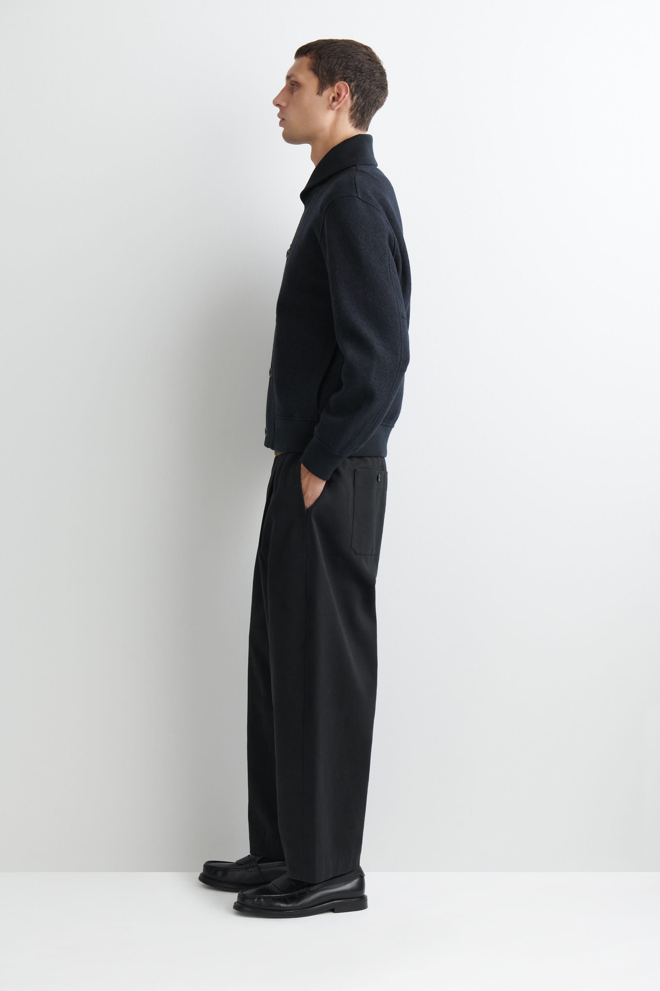 A navy boiled-wool shawl-collar jacket is shown in left side profile, featuring a structured shawl collar, ribbed cuffs, and a ribbed hem. The jacket sits at the waist to highlight its clean, tailored fit and is styled with wide-leg black trousers and black leather loafers. A navy boiled-wool shawl-collar jacket is shown in left side profile, featuring a structured shawl collar, ribbed cuffs, and a ribbed hem. The jacket sits at the waist to highlight its clean, tailored fit and is styled with wide-leg black trousers and black leather loafers.