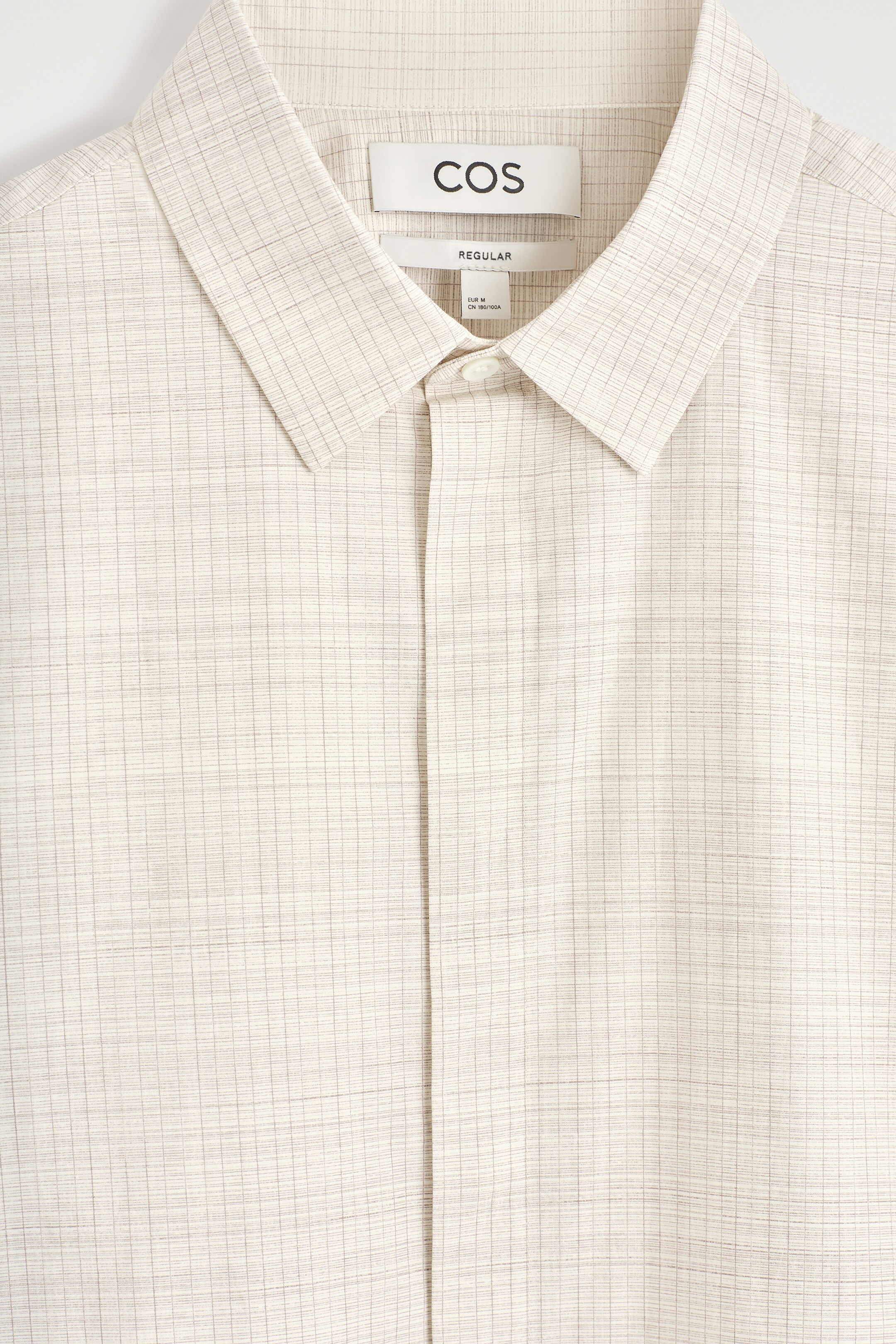 Agrandir l'image: Le devant d'une chemise beige clair présente un motif à carreaux subtil et fin. Elle est dotée d'un col pointu et d'un bouton en nacre visible en haut de sa patte de boutonnage.