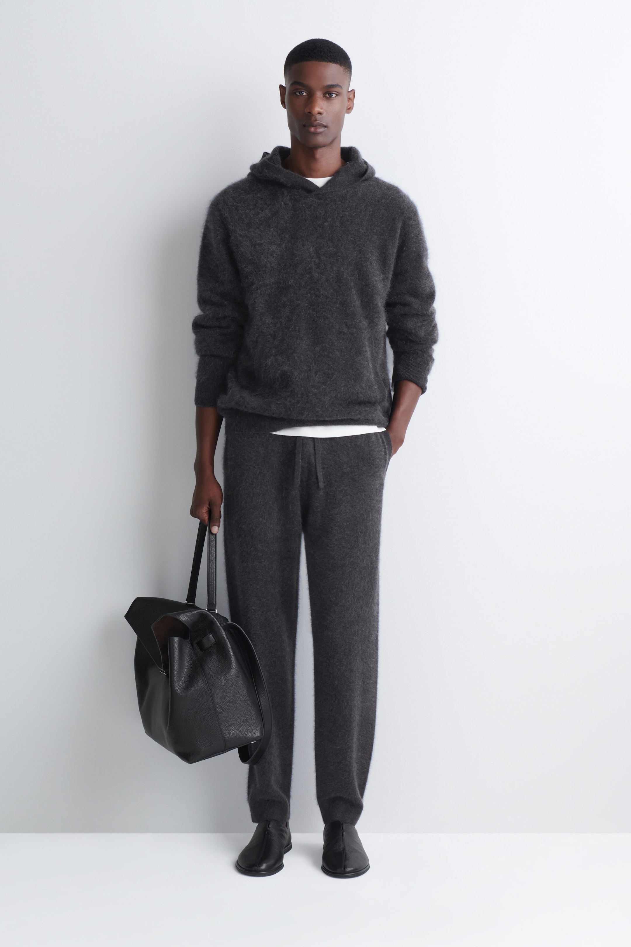 A dark gray brushed cashmere hoodie with a soft, textured finish is worn by the model, layered over a white T-shirt and paired with matching knit trousers. The model is carrying a black leather bag in one hand.