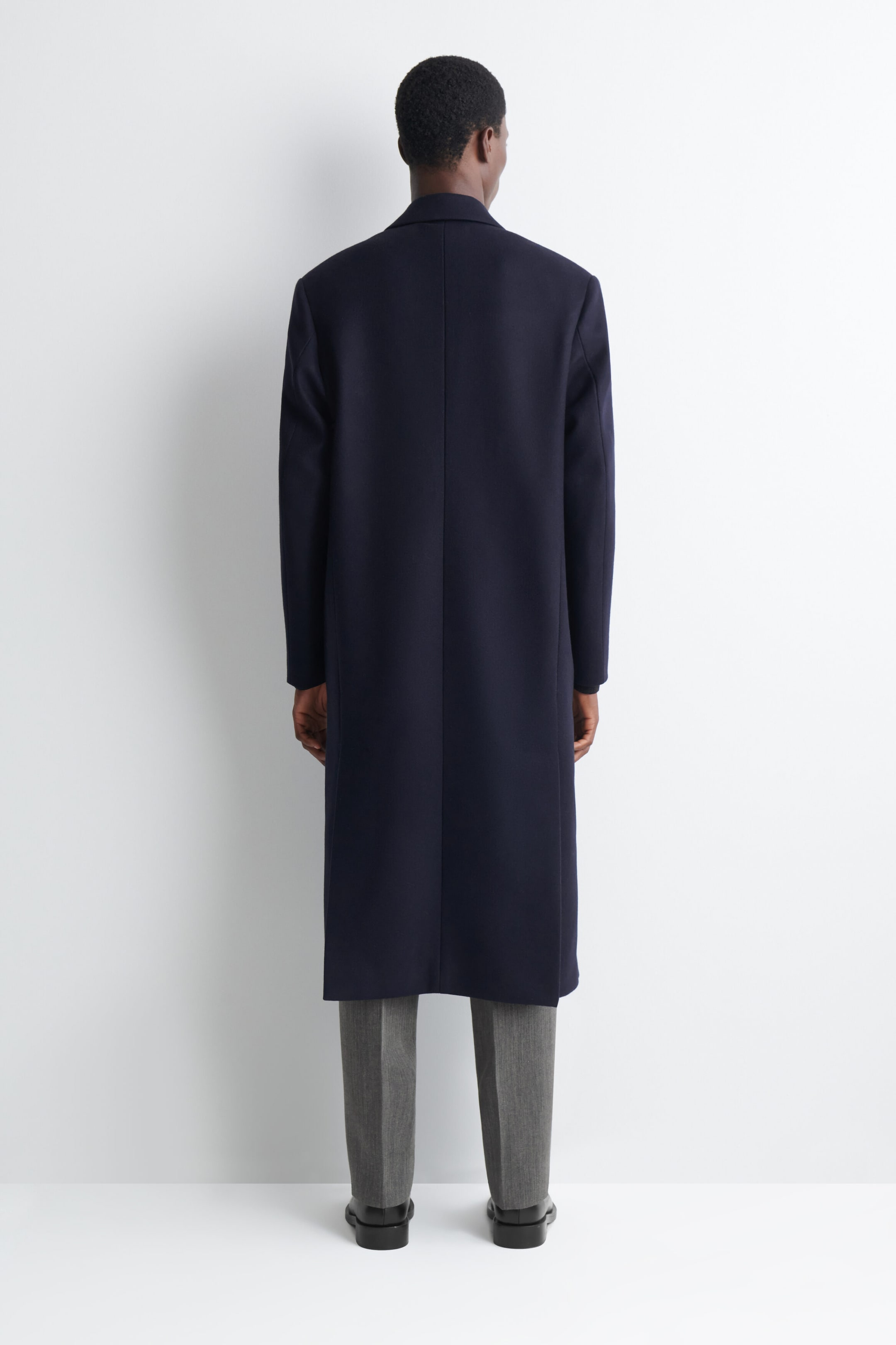The model is wearing a navy long coat, featuring a clean back design and structured shoulders. The coat falls below the knees and is styled with grey trousers and black shoes. The model is wearing a navy long coat, featuring a clean back design and structured shoulders. The coat falls below the knees and is styled with grey trousers and black shoes.