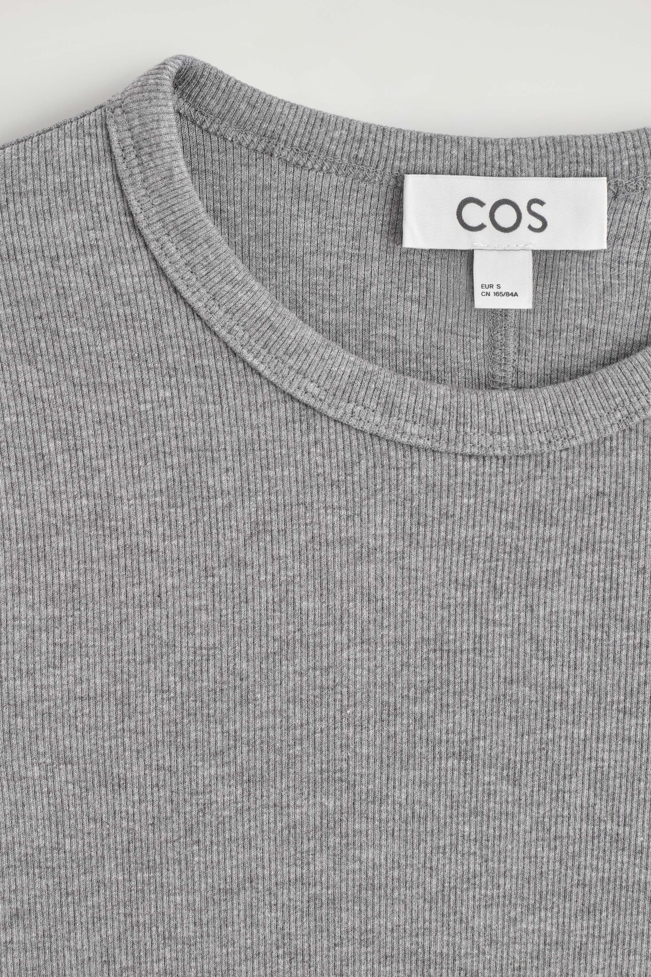 The slim ribbed cotton long-sleeved top is shown in a close-up, front-facing view, showcasing its round crew neckline and fine-ribbed grey mélange texture. A white brand label is stitched inside the back collar, displaying the brand name "COS". The slim ribbed cotton long-sleeved top is shown in a close-up, front-facing view, showcasing its round crew neckline and fine-ribbed grey mélange texture. A white brand label is stitched inside the back collar, displaying the brand name "COS".
