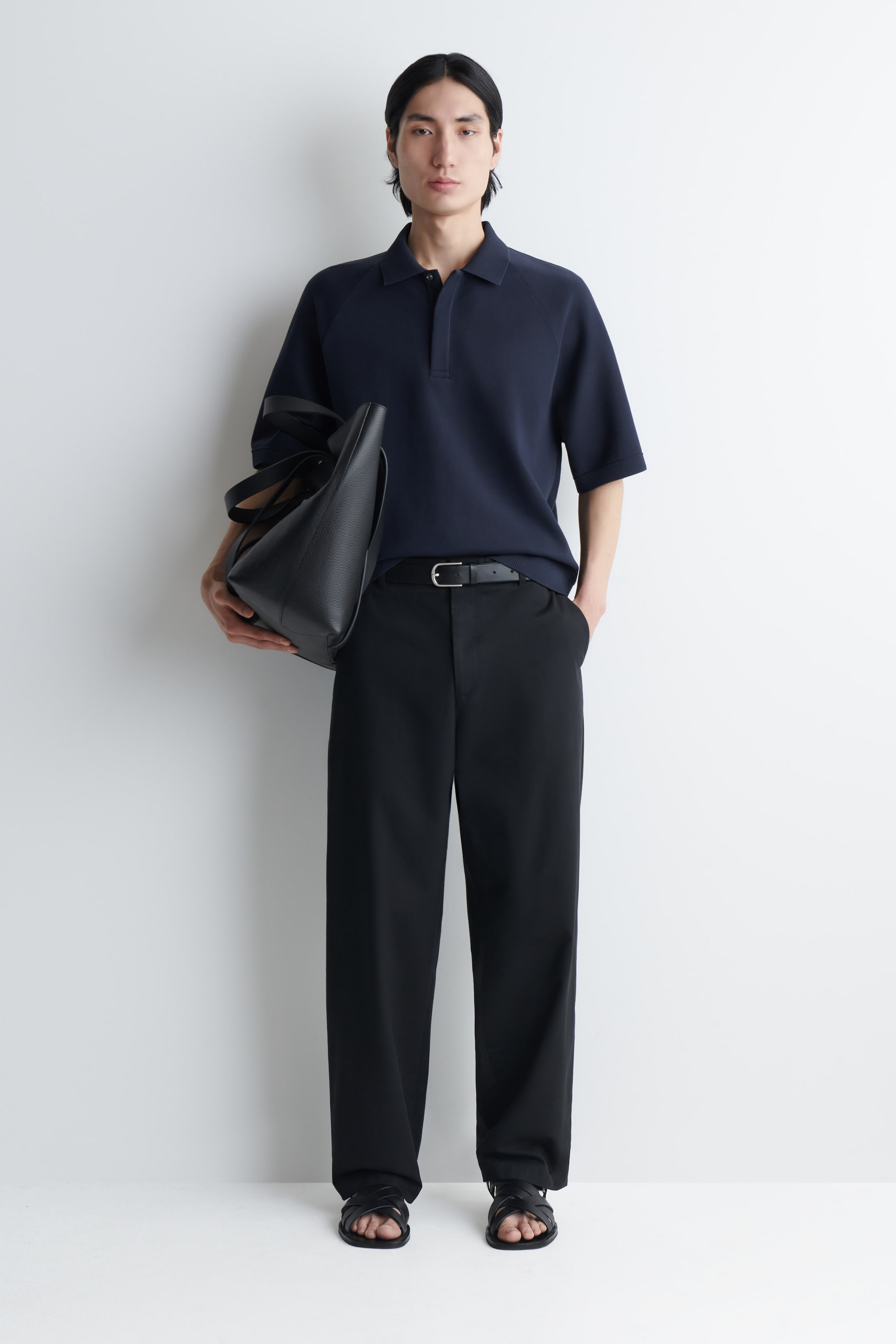 Grotere afbeelding bekijken: Een marineblauw polohemd in scuba-jersey, met een gestructureerde kraag, korte raglanmouwen en een nette knopenlijst, wordt gedragen door het model. Het hemd is gestyled met een zwarte wijde broek, een zwarte leren riem en een zwarte leren tote bag die onder één arm wordt gedragen.