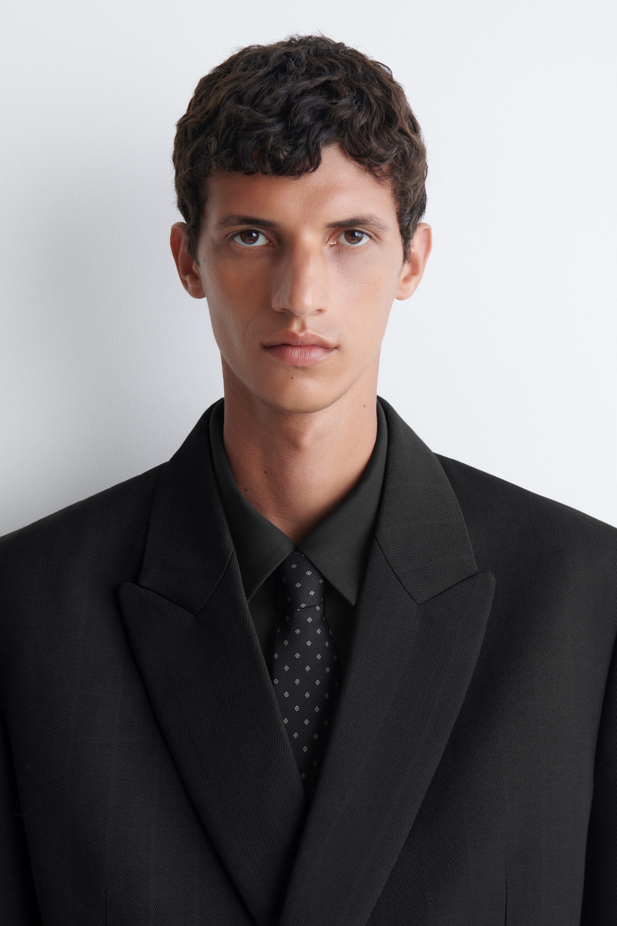 A dark silk tie featuring a small, light-coloured geometric foulard pattern is being worn by the model. The tie is styled under a black collared shirt and a matching black tailored blazer. A dark silk tie featuring a small, light-coloured geometric foulard pattern is being worn by the model. The tie is styled under a black collared shirt and a matching black tailored blazer.