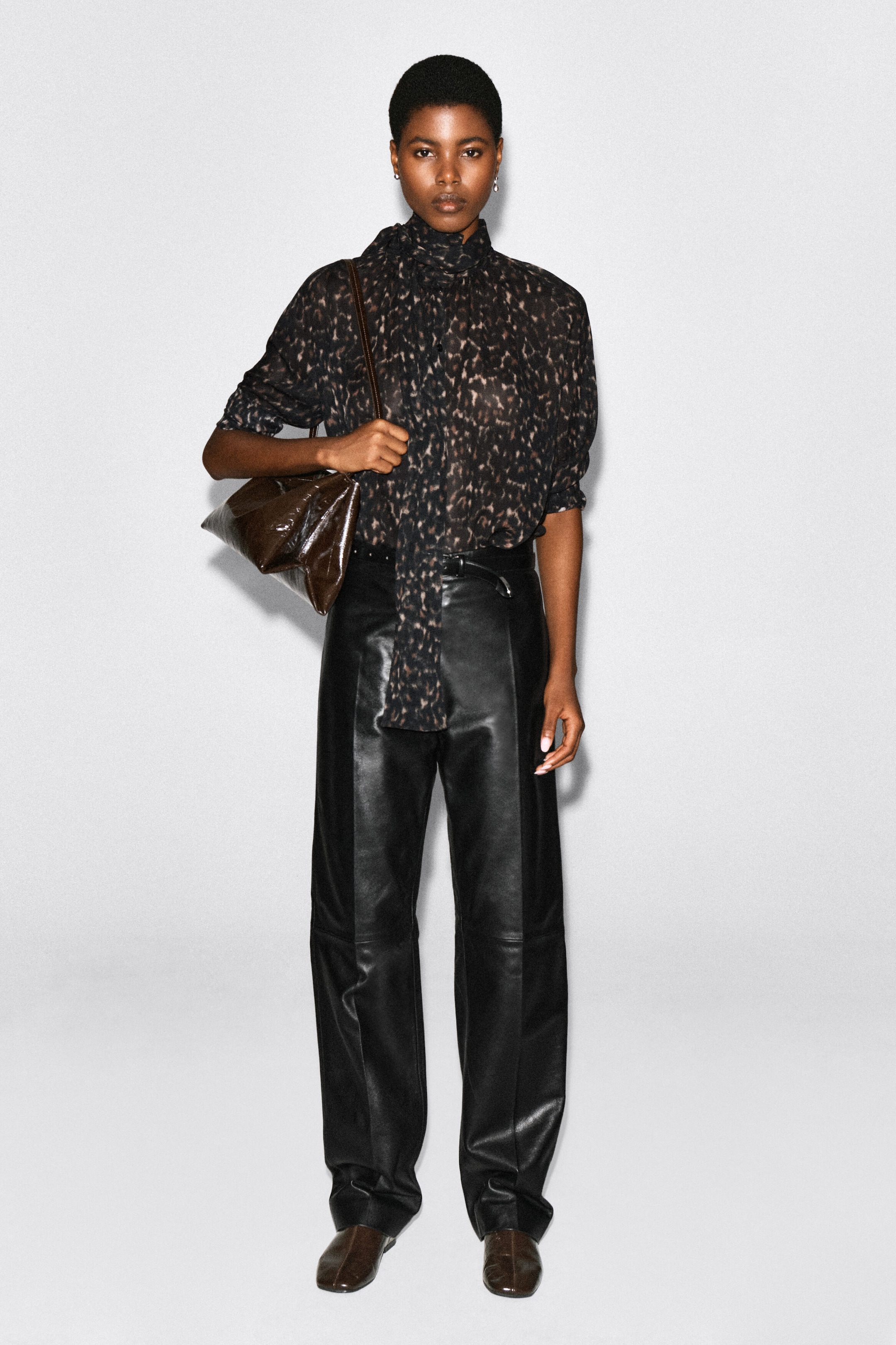 A leopard-print fluid scarf-detail top in a dark, semi-sheer fabric is worn by the model, tucked into black leather trousers. The top features a high collar with an integrated long matching scarf draped down the front, and is styled with a dark brown shoulder bag. A leopard-print fluid scarf-detail top in a dark, semi-sheer fabric is worn by the model, tucked into black leather trousers. The top features a high collar with an integrated long matching scarf draped down the front, and is styled with a dark brown shoulder bag.
