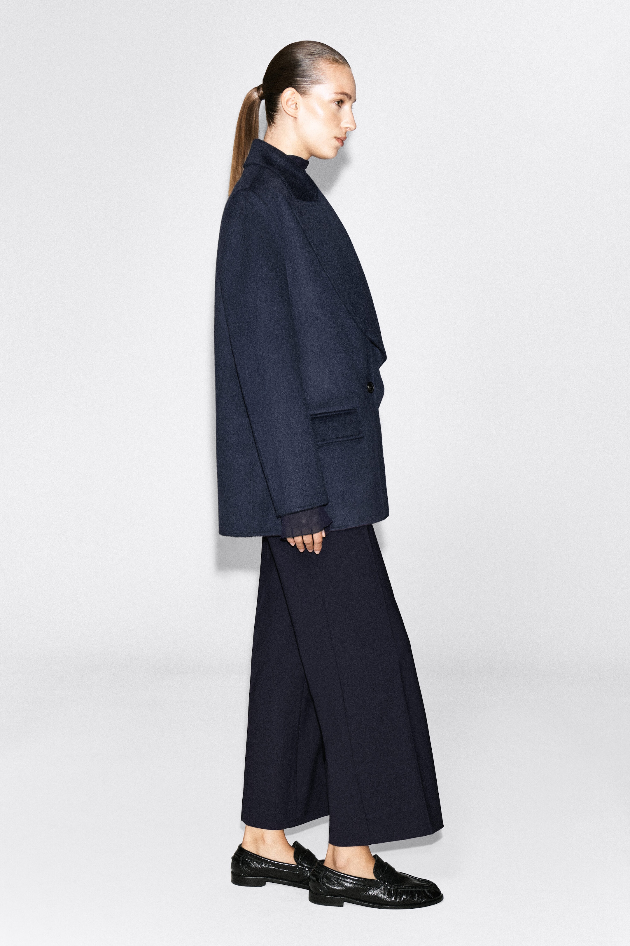 A dark navy blue wool-blend blazer is worn by the model, who is standing in profile to showcase its structured, relaxed fit and exaggerated lapels. The blazer is paired with matching wide-leg trousers and black leather loafers.
