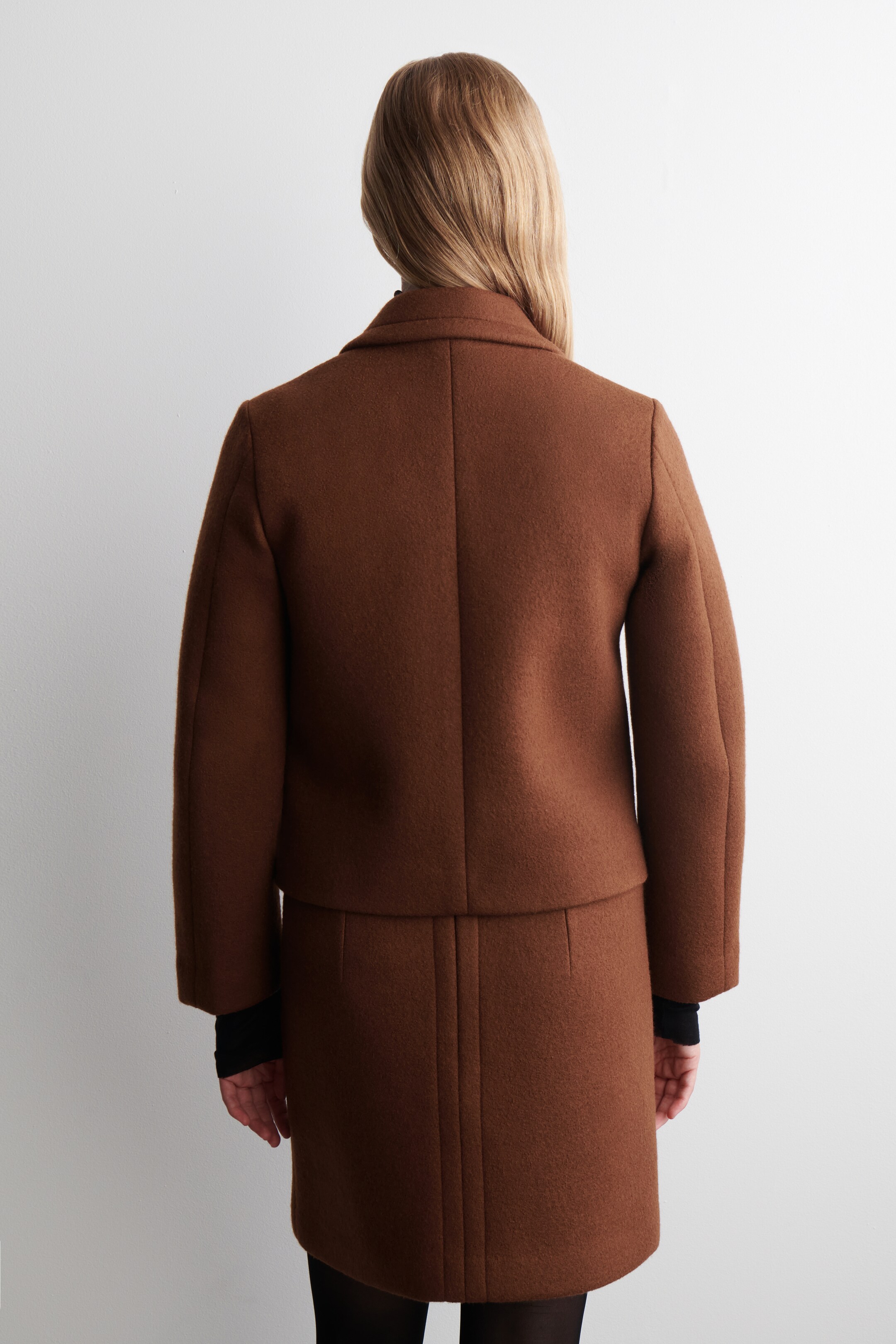 View larger image: A brown cropped jacket and matching mini skirt are being presented from the back. The jacket is featuring a collar at the back of the neck and long sleeves, worn over a black top, while the skirt is showing vertical seam details and is styled over black tights.