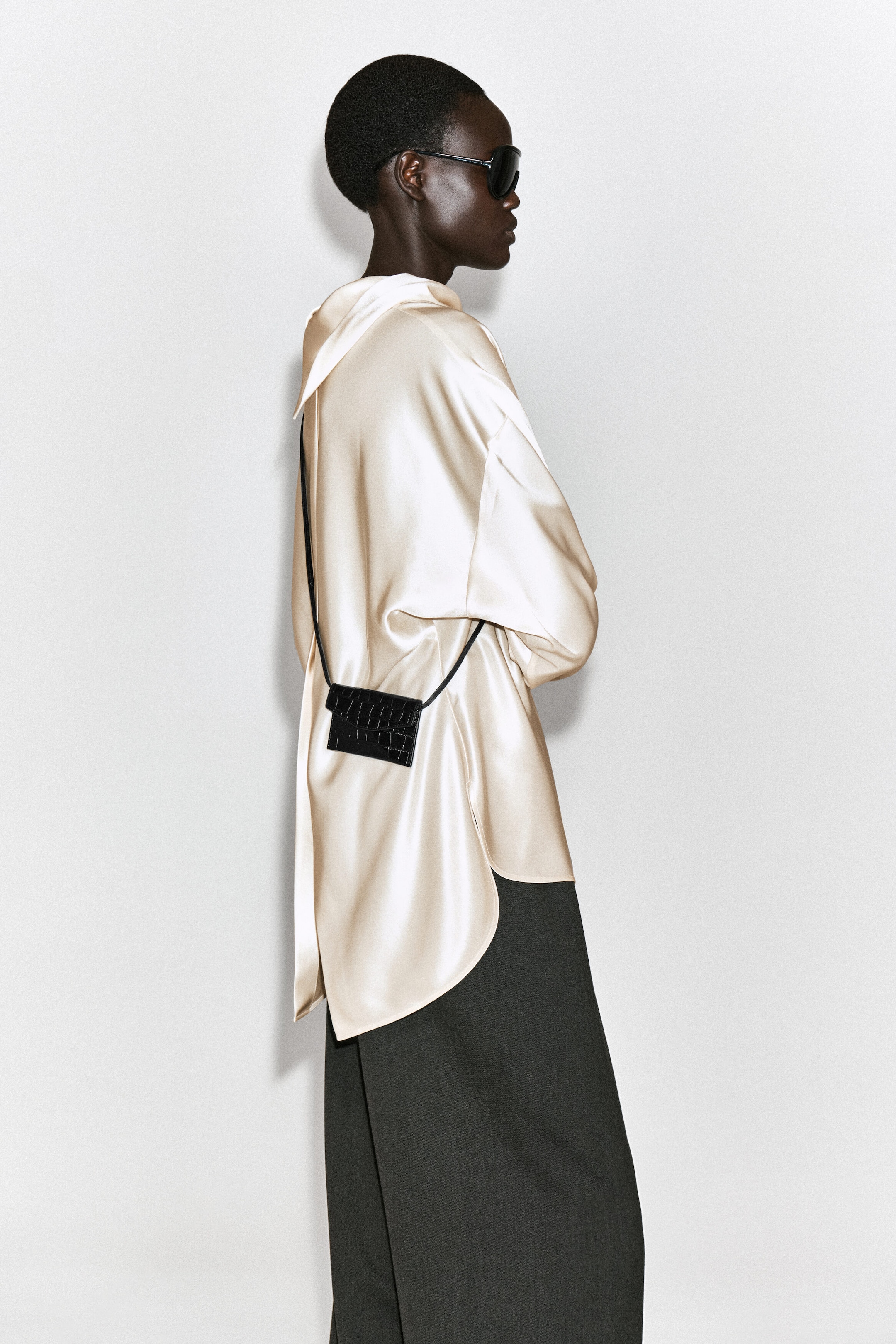 A black leather card holder with a crocodile-embossed texture is worn crossbody with a thin strap, resting on the hip of a model facing right. The rectangular card holder features a fold-over flap closure and is styled over a cream satin blouse. A black leather card holder with a crocodile-embossed texture is worn crossbody with a thin strap, resting on the hip of a model facing right. The rectangular card holder features a fold-over flap closure and is styled over a cream satin blouse.