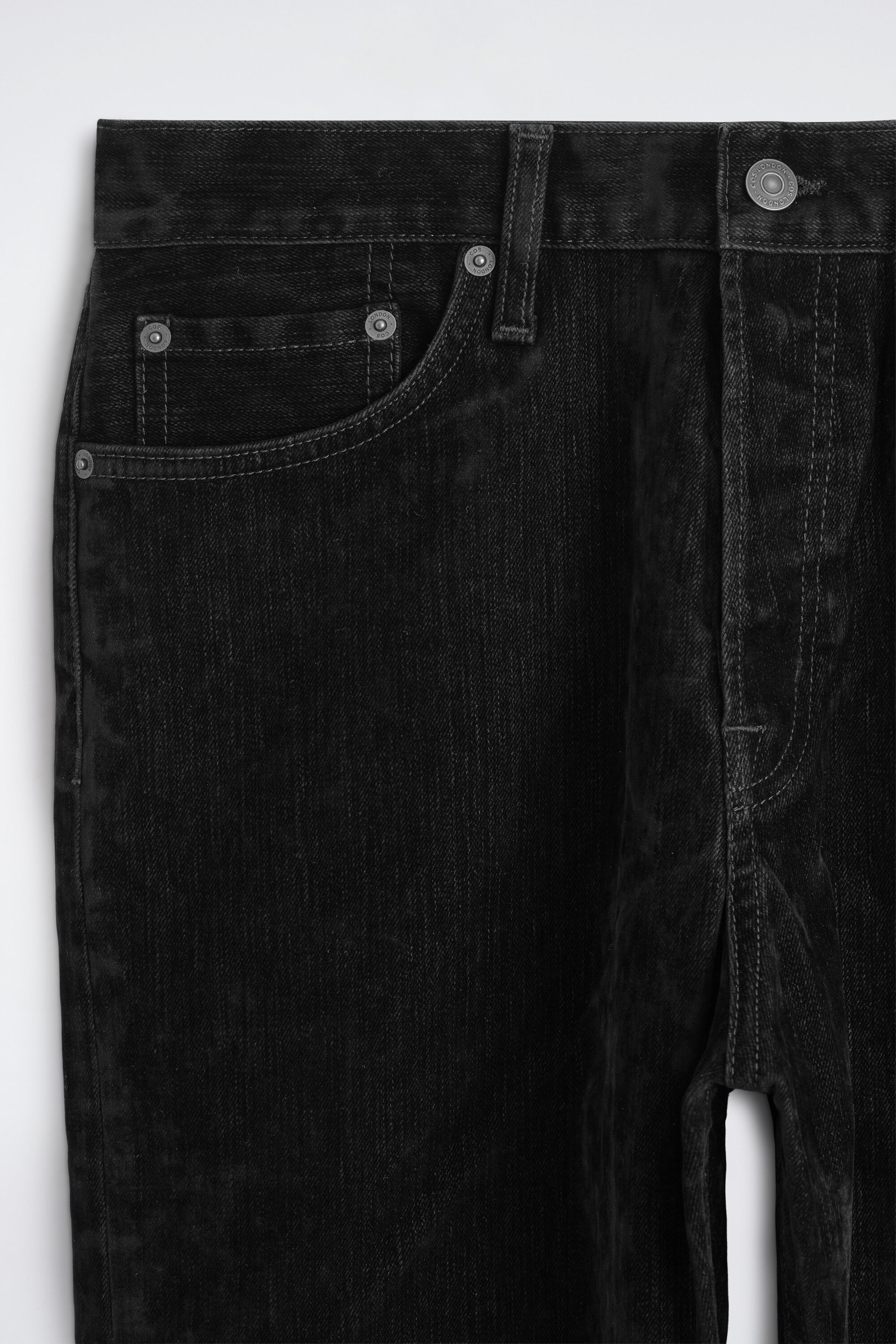 Los Signature Flocked Straight-Leg Jeans muestran una vista frontal en primer plano de la cintura y la parte superior de la cadera, destacando el tejido vaquero flocado en negro oscuro con un acabado suave y texturizado. El diseño presenta una cintura clásica con trabillas para cinturón, cierre de botón metálico plateado tipo shank y un bolsillo delantero curvado con remaches metálicos a juego.