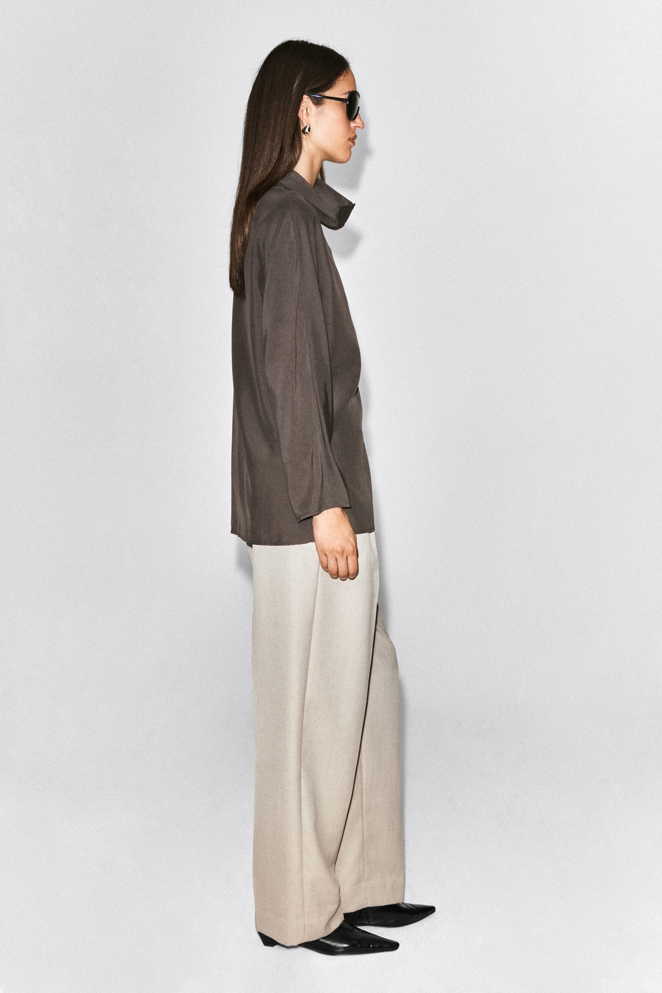 The model is wearing light beige straight-leg trousers with a relaxed silhouette and front pleats, gently pooling over black pointed-toe shoes. The trousers are styled with a dark grey long-sleeved high-neck top. The model is wearing light beige straight-leg trousers with a relaxed silhouette and front pleats, gently pooling over black pointed-toe shoes. The trousers are styled with a dark grey long-sleeved high-neck top.
