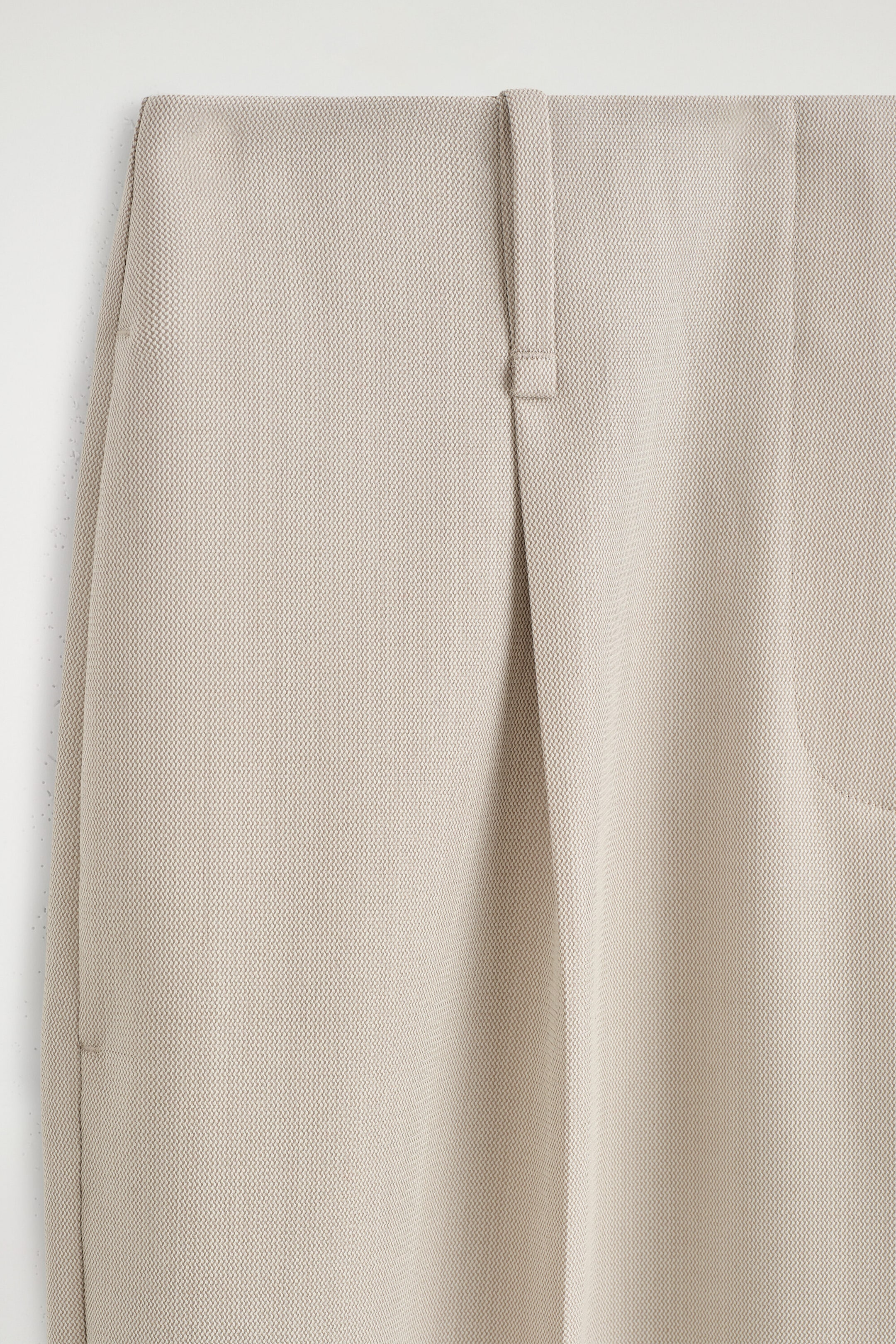 This close-up front view of light beige textured trousers showcases a structured waistband with a single vertical belt loop and a prominent front pleat. The finely woven fabric drapes downward, highlighting a subtle textured pattern. This close-up front view of light beige textured trousers showcases a structured waistband with a single vertical belt loop and a prominent front pleat. The finely woven fabric drapes downward, highlighting a subtle textured pattern.
