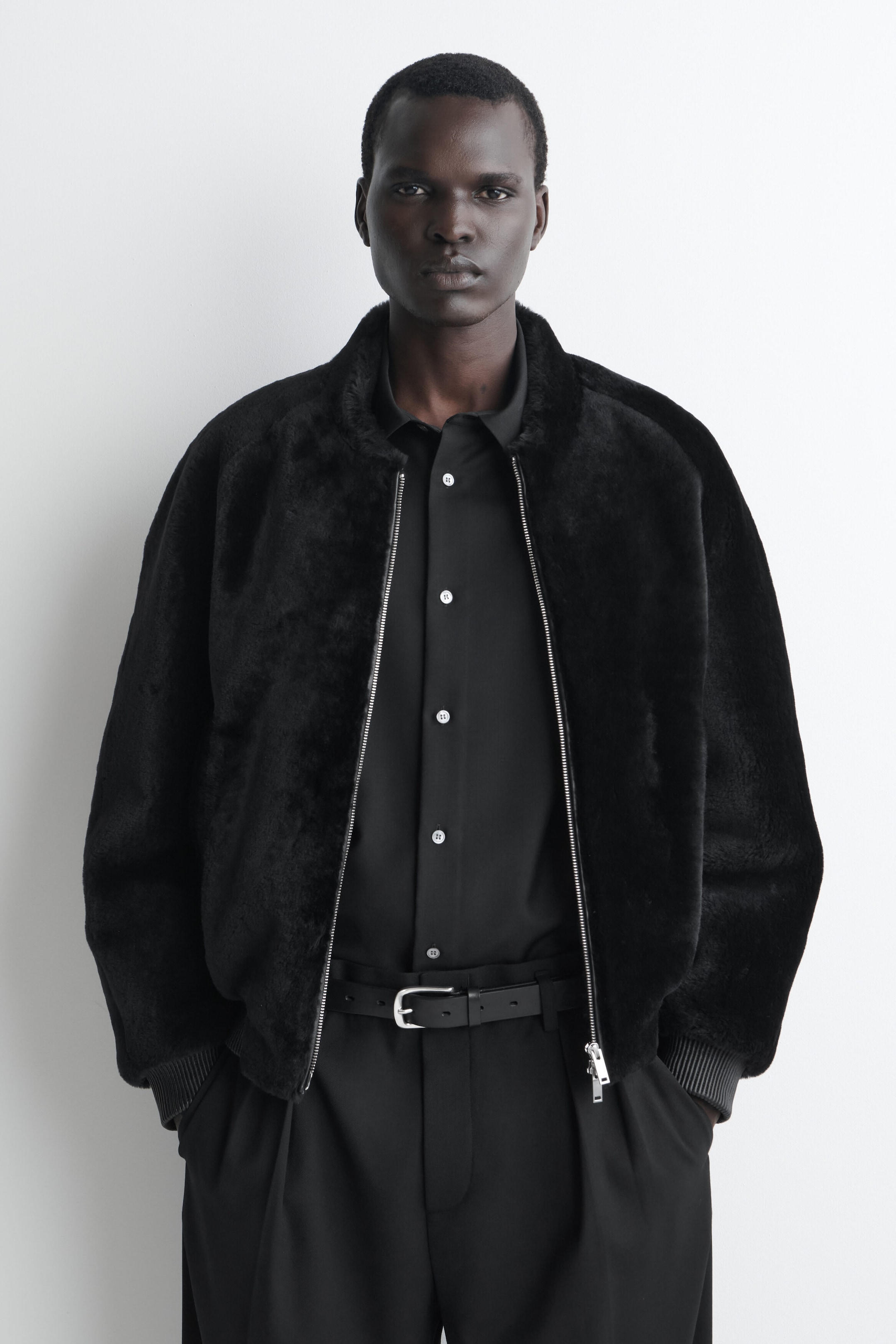 A padded shearling bomber jacket is shown from the front, highlighting its plush black texture, silver front zipper, and ribbed cuffs. The jacket is worn open over a black button-down shirt and black trousers.