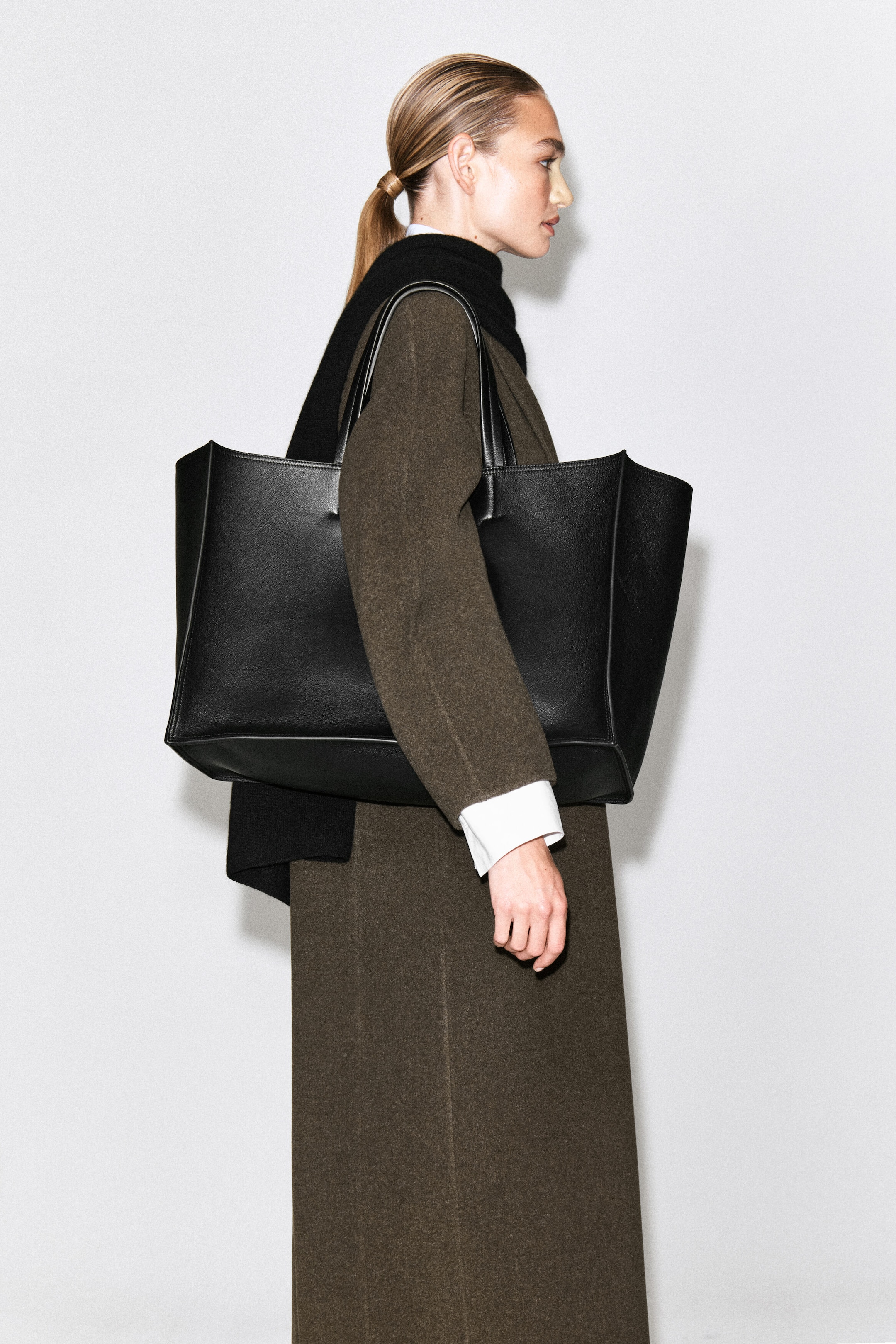 A large black leather tote bag is being carried on the model’s shoulder, who is wearing a dark olive-brown wool coat and a black scarf. A large black leather tote bag is being carried on the model’s shoulder, who is wearing a dark olive-brown wool coat and a black scarf.