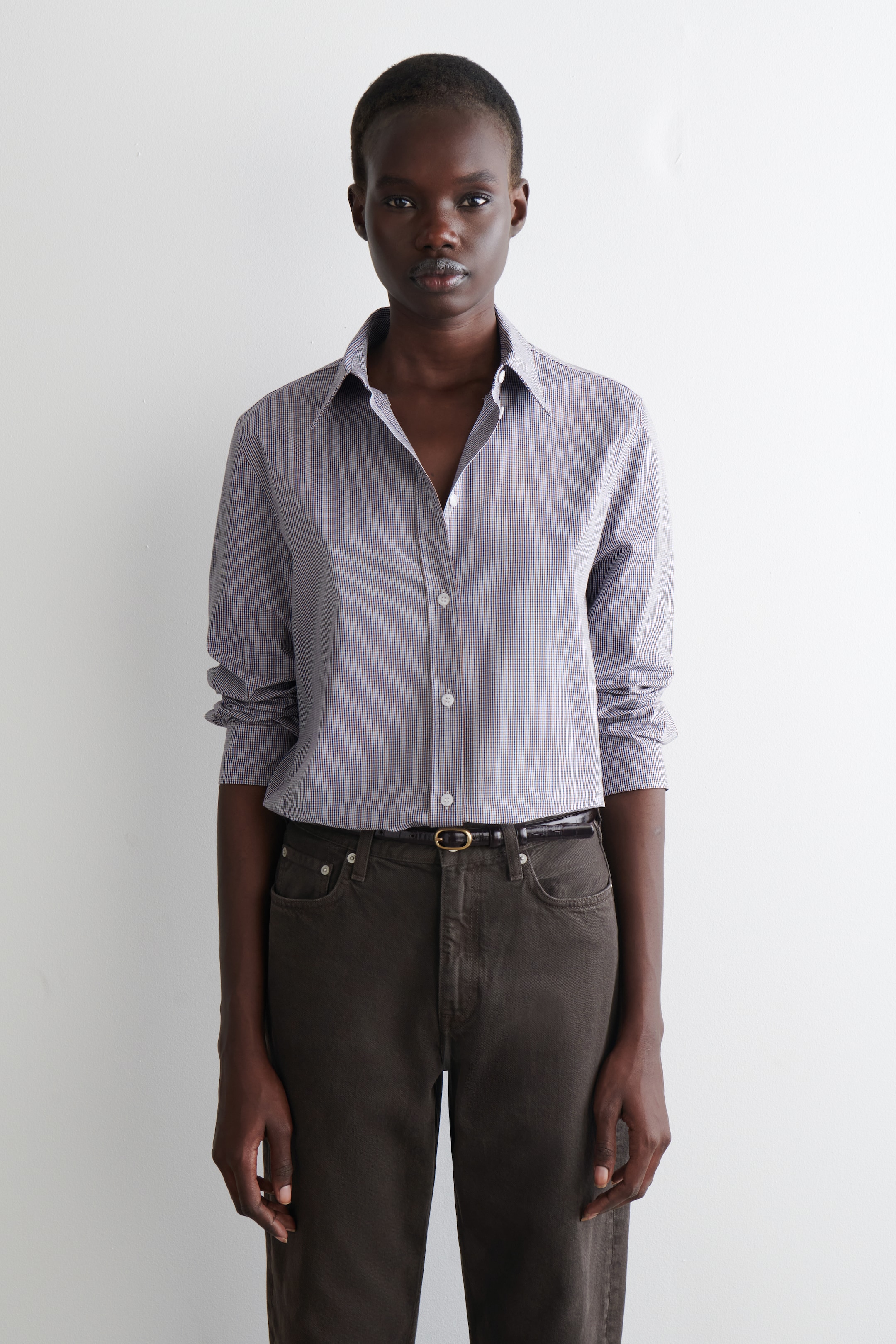Visualizza immagine più grande: Una camicia a quadretti micro di colore grigio chiaro è presentata, caratterizzata da un colletto a punta, un'abbottonatura completa con bottoni bianchi e maniche lunghe arrotolate. La camicia è indossata infilata in pantaloni marrone scuro, assicurata da una cintura scura con una fibbia ovale dorata, con tutti gli articoli rivolti in avanti.