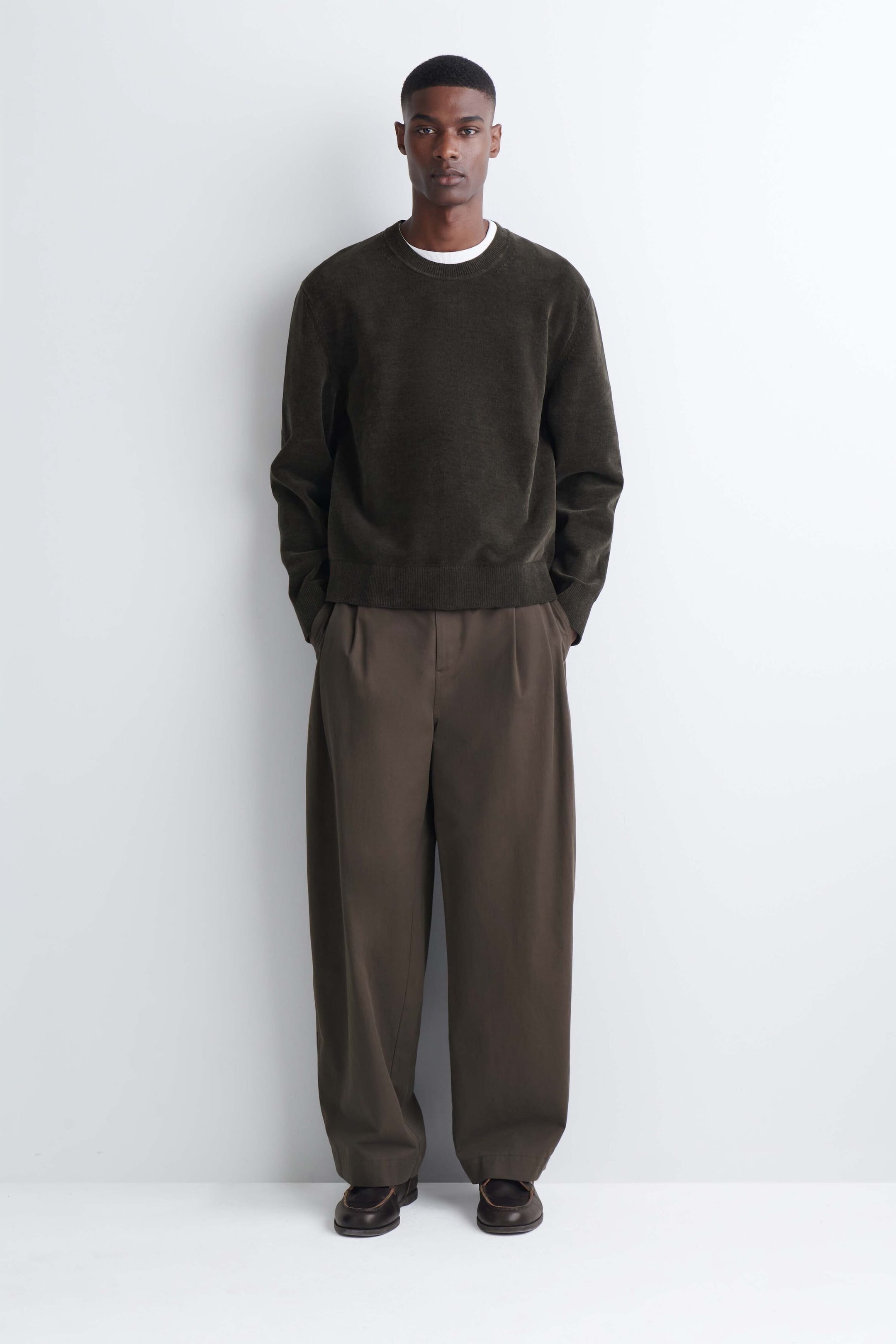 A dark green cotton-chenille crew neck jumper is worn by the model, layered over a white T-shirt and paired with wide-leg brown trousers. The jumper features a soft, textured finish with ribbed detailing on the collar, cuffs, and hem. A dark green cotton-chenille crew neck jumper is worn by the model, layered over a white T-shirt and paired with wide-leg brown trousers. The jumper features a soft, textured finish with ribbed detailing on the collar, cuffs, and hem.