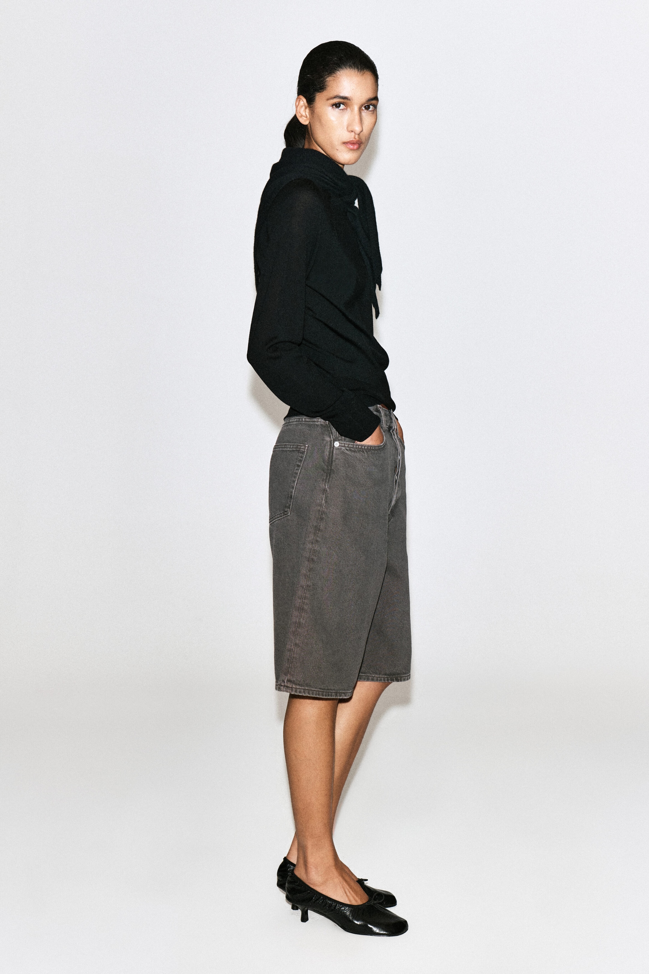 Longline denim shorts in a washed espresso hue are shown from a side profile facing right, highlighting their high-rise waist and classic five-pocket design. The knee-length shorts feature tonal topstitching and are styled with a tucked-in black long-sleeve top to emphasize their structured silhouette. Longline denim shorts in a washed espresso hue are shown from a side profile facing right, highlighting their high-rise waist and classic five-pocket design. The knee-length shorts feature tonal topstitching and are styled with a tucked-in black long-sleeve top to emphasize their structured silhouette.