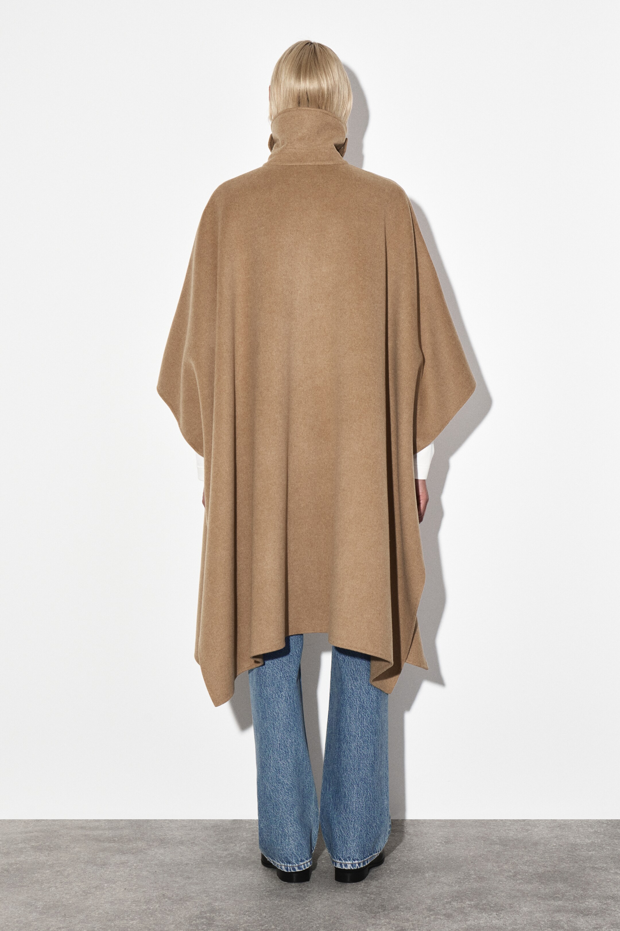 Agrandir l'image: Cape en laine double face à longue ligne, présentée comme DOS. Cette cape de couleur camel présente un col cheminée haut et une silhouette large et fluide, tombant librement des épaules. Ses côtés ouverts révèlent un vêtement blanc à manches longues en dessous, et son ourlet s'étend jusqu'au milieu de la cuisse.