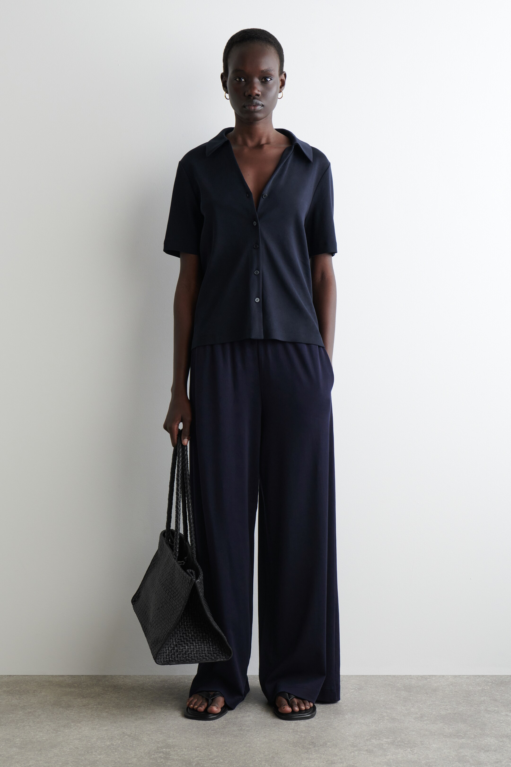 Agrandir l'image: Une chemise à manches courtes bleu marine foncé avec un col ouvert en V et des boutons sur le devant est assortie à un pantalon large, formant un ensemble décontracté. Un sac fourre-tout noir tissé avec des poignées tressées est tenu, et des sandales noires à lanières sont portées, complétant l'ensemble.