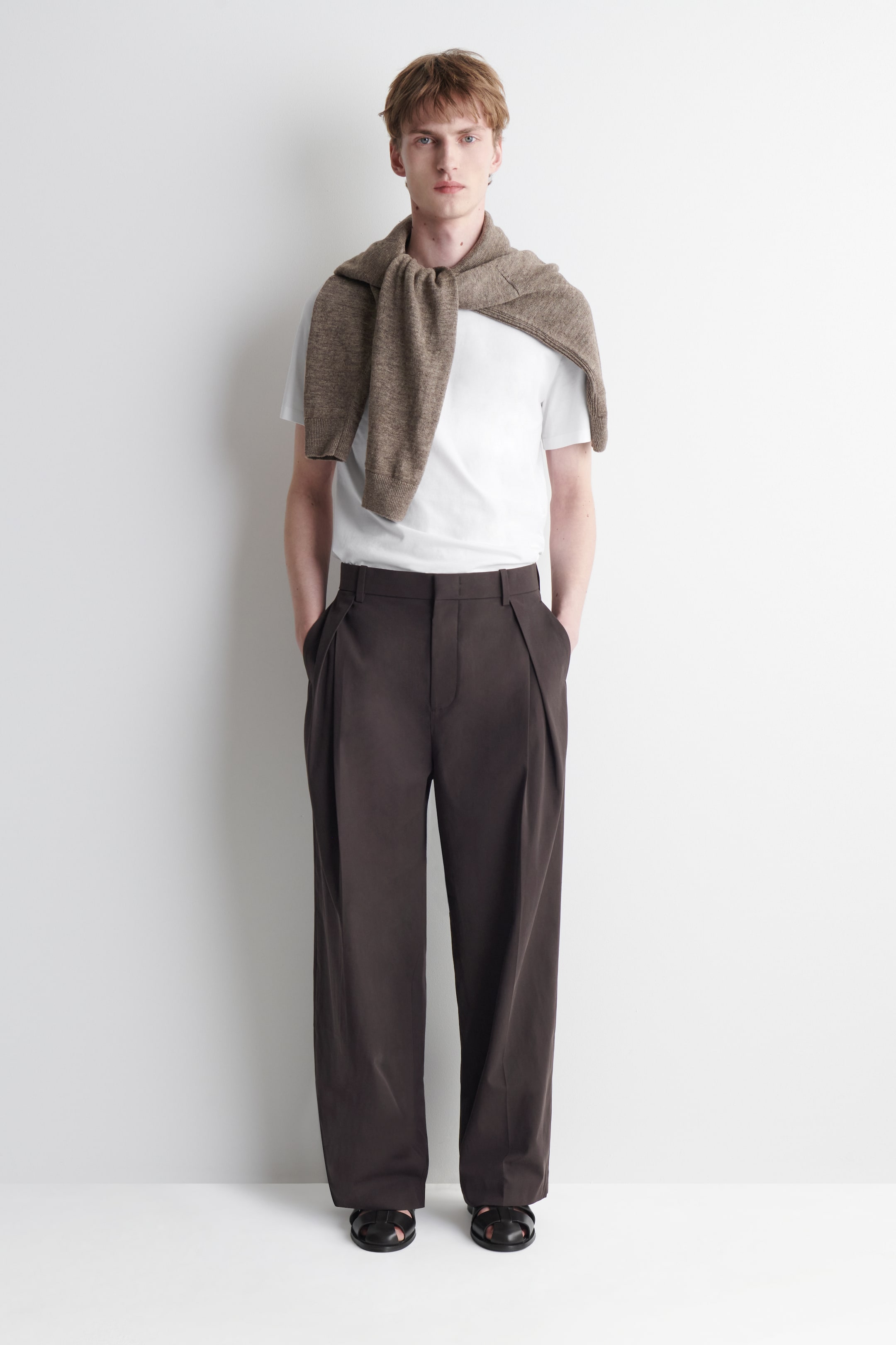 View larger image: Pleated cotton relaxed-fit wide-leg trousers in dark brown are worn by the model, whose hands are placed in the side pockets. The trousers feature deep front pleats and a loose silhouette, and are styled with a tucked-in white T-shirt and a brown knit sweater draped over the shoulders.