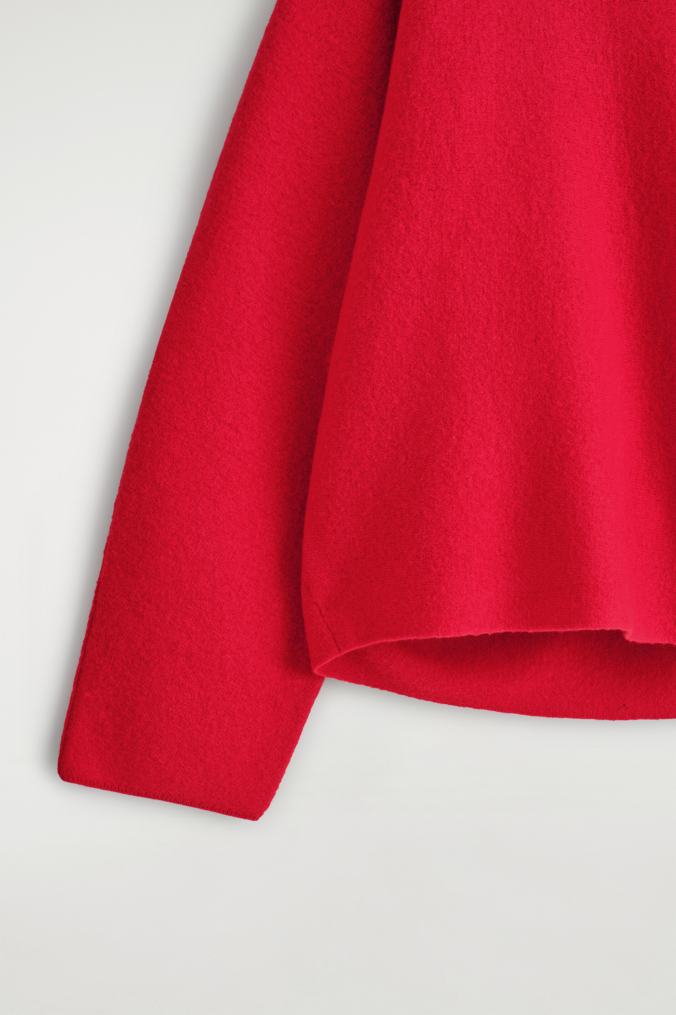 Agrandir l'image: Un gros plan montre une partie d'un pull rouge vif, avec une texture bouillie sur son tissu. Le corps du vêtement présente un ourlet subtilement arrondi, avec une manche apparaissant à gauche.