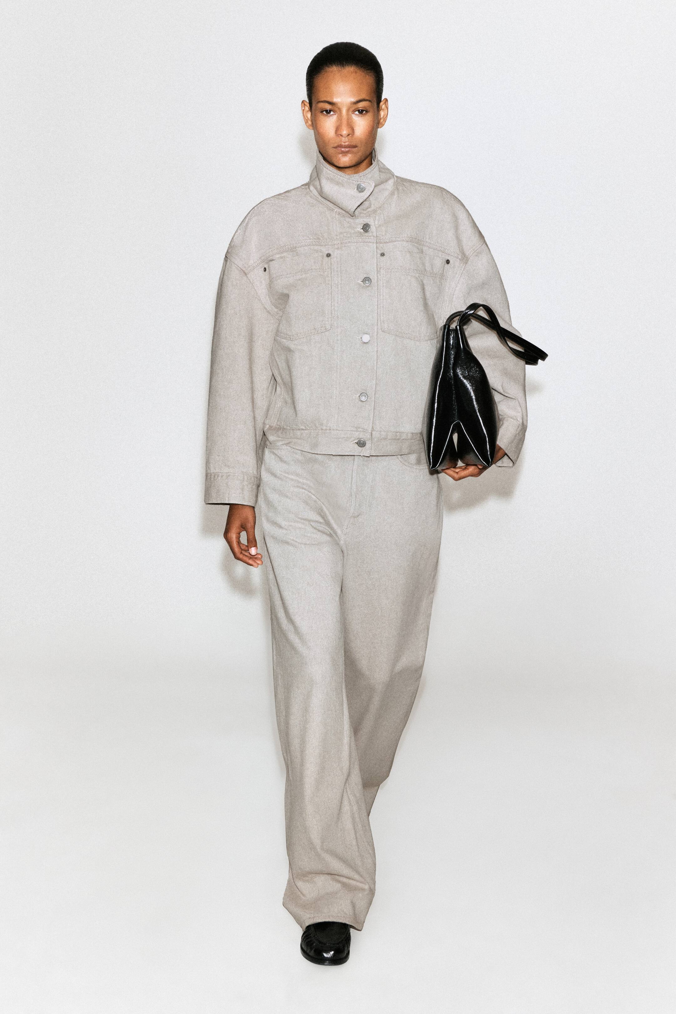 Grösseres Bild ansehen: Ein hellgraue Jeansjacke mit hohem Kragen und dazu passende weite Hosen werden vom Model getragen. Das Model hält eine schwarze Handtasche aus Lackleder mit strukturierter Form und trägt schwarze Loafer.