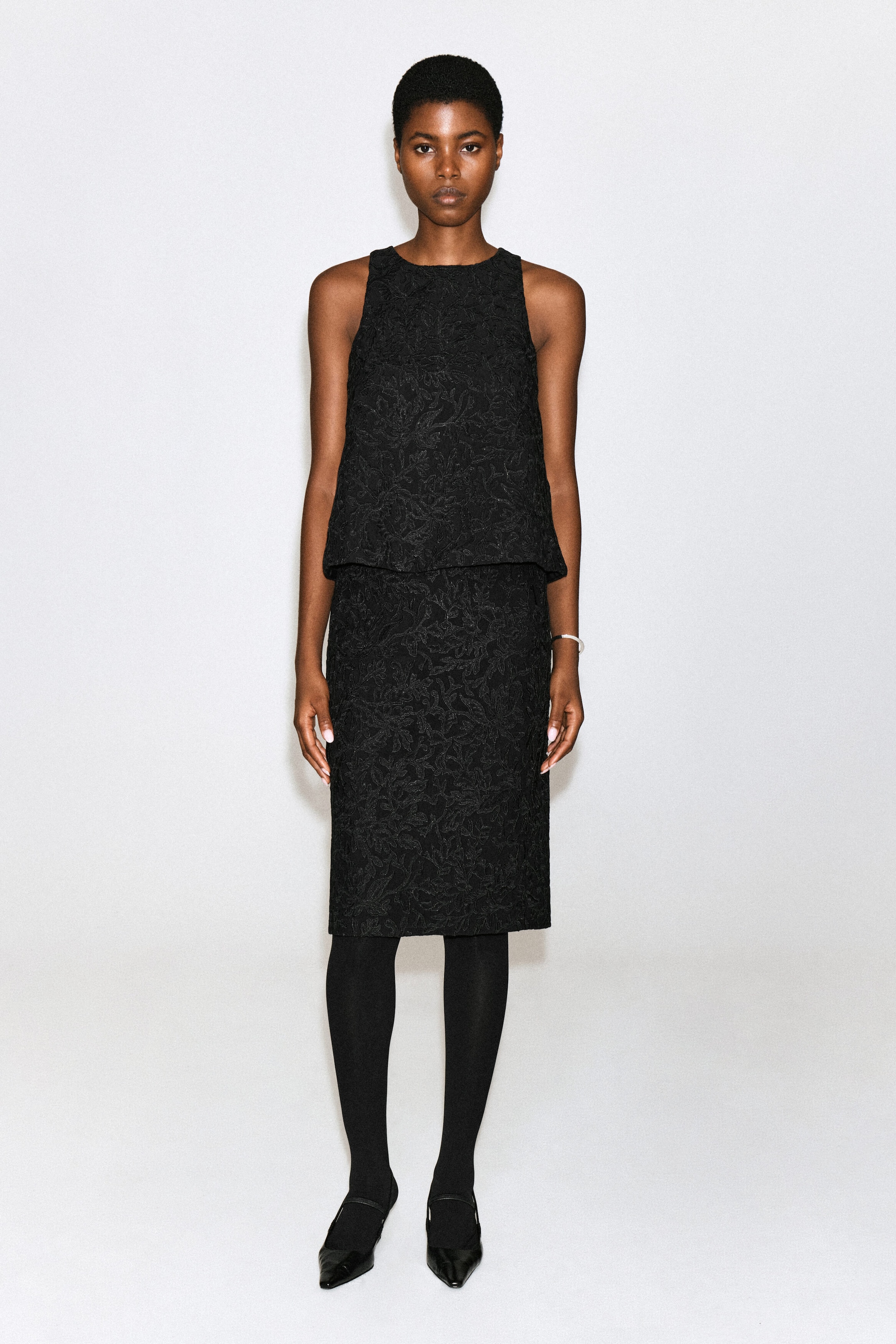 Un haut noir sans manches orné d’une broderie florale en dentelle texturée est porté par le mannequin. Ce haut à coupe décontractée est agencé avec une jupe en dentelle assortie longueur genou, des collants noirs et des chaussures noires à bout pointu.