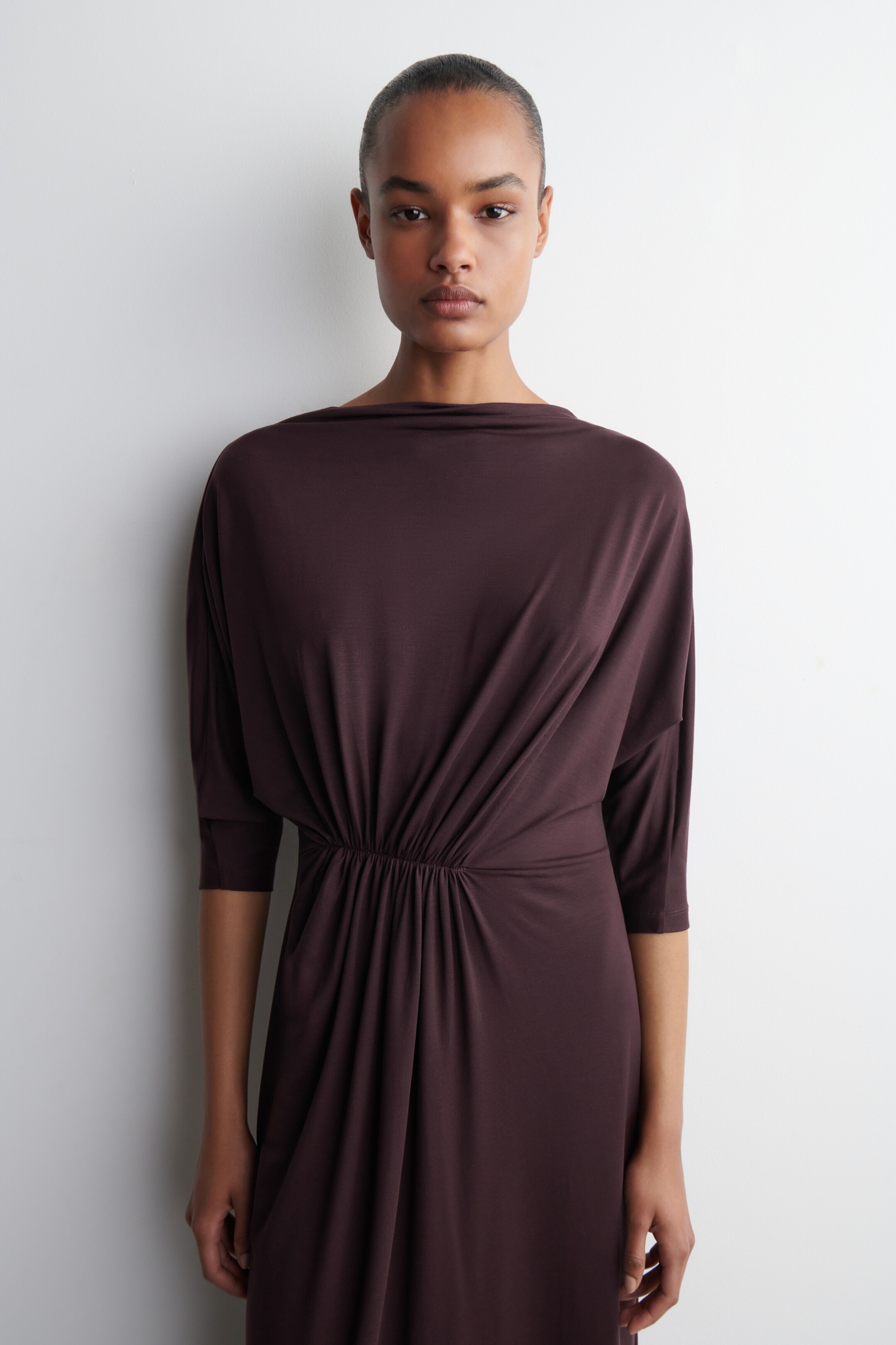 Agrandir l'image: Une robe prune-brun foncé présente un col bateau drapé et des manches dolman trois-quarts. Le tissu se rassemble à une taille élastiquée, d'où la jupe s'écoule vers le bas.