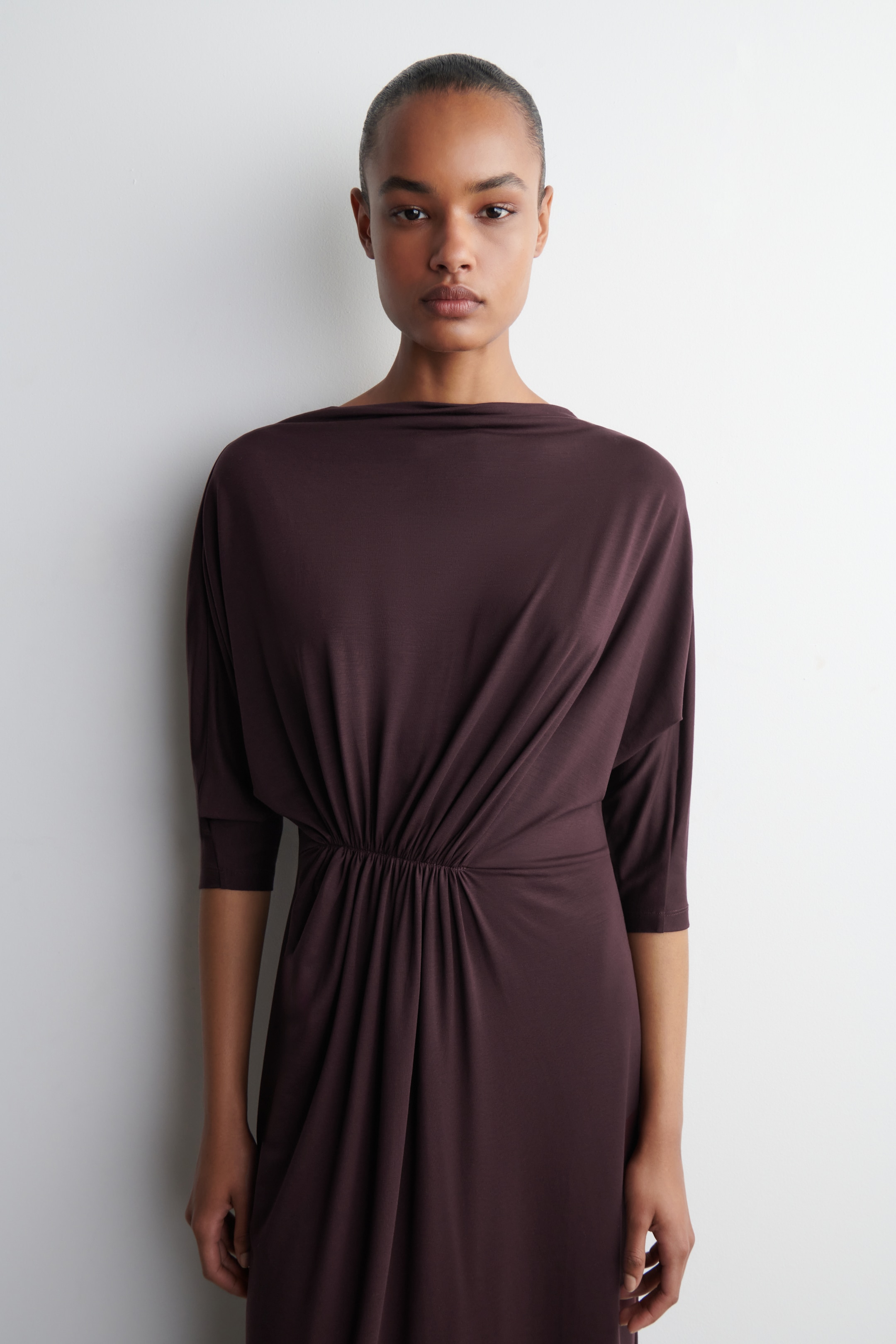 Größeres Bild anzeigen: Ein tiefes pflaumenbraunes Kleid zeichnet sich durch einen drapierten Bootsausschnitt und dreiviertellange Dolman-Ärmel aus. Der Stoff sammelt sich an einer elastischen Taille, von der der Rock nach unten fließt.