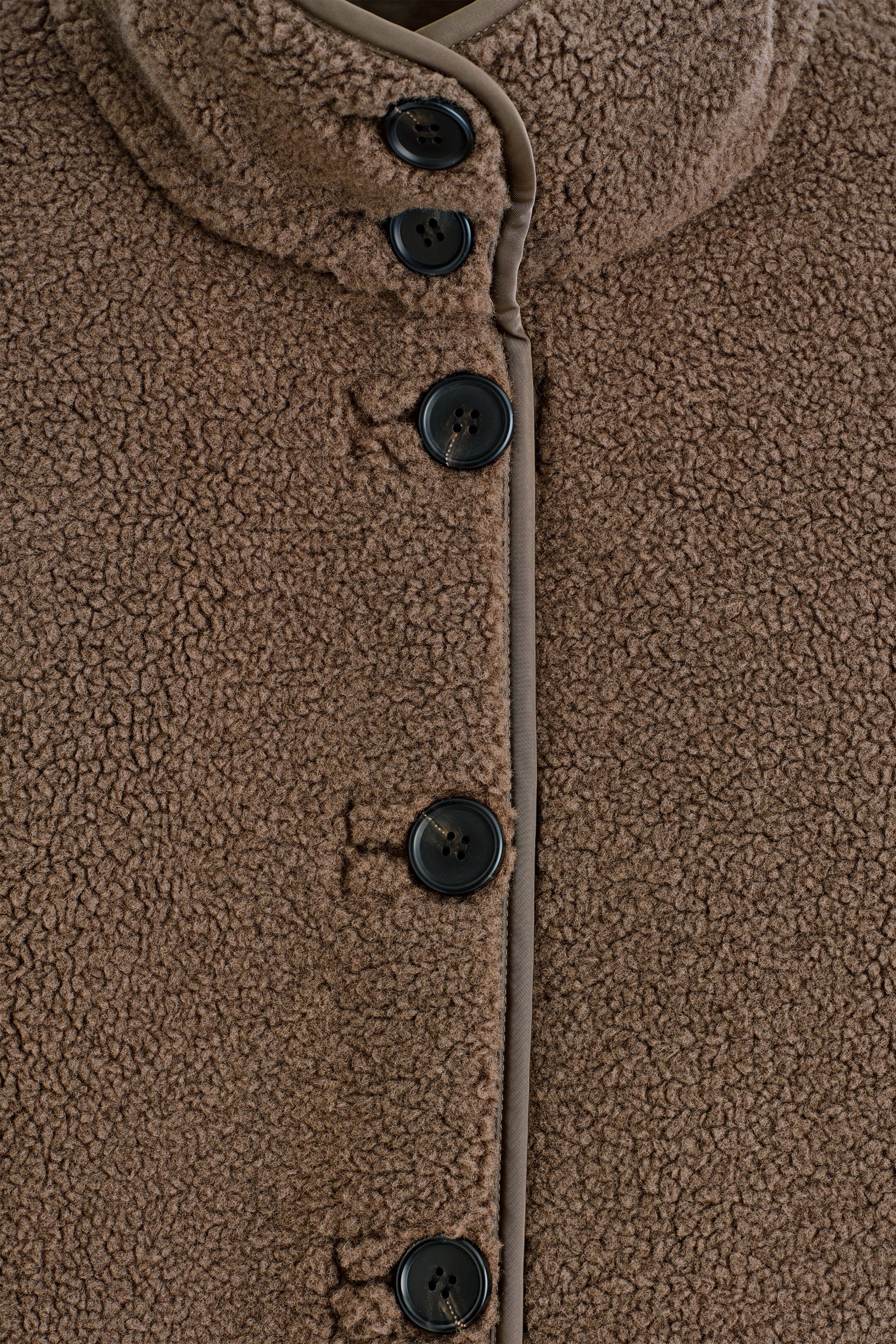 Agrandir l'image: Une veste en polaire teddy marron est présentée de face, avec une fermeture à boutons. Des boutons ronds foncés à quatre trous ferment la veste, tandis qu'un liseré marron foncé souligne la patte de boutonnage et le bord du col montant.