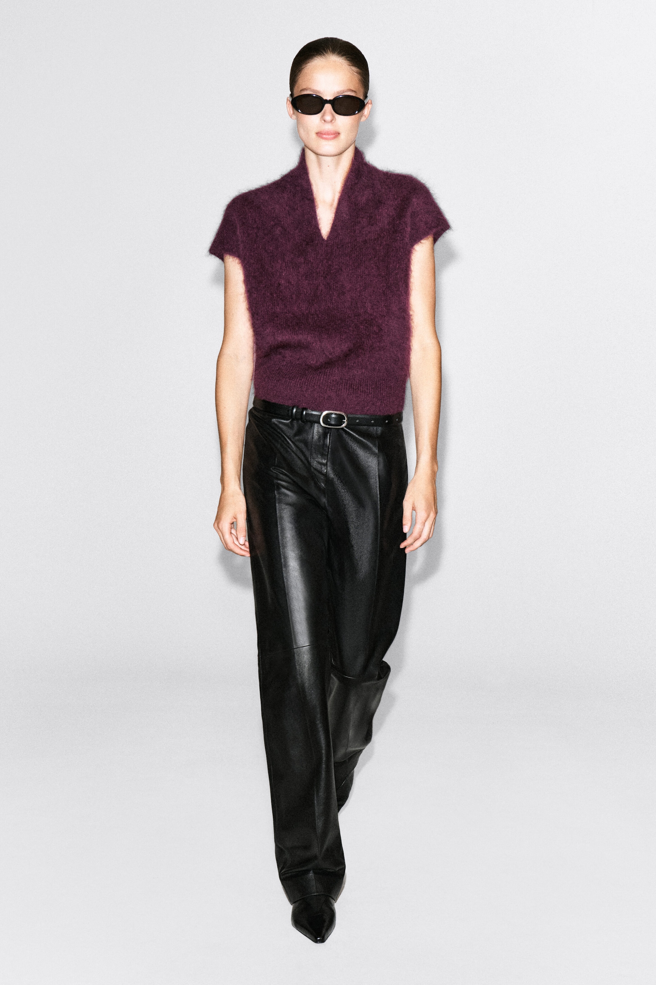 The draped brushed merino wool top in a deep plum color is being worn by the model, featuring a fuzzy texture, a V-neckline, and short cap sleeves. The top is tucked into wide-leg black leather trousers styled with a black leather belt.