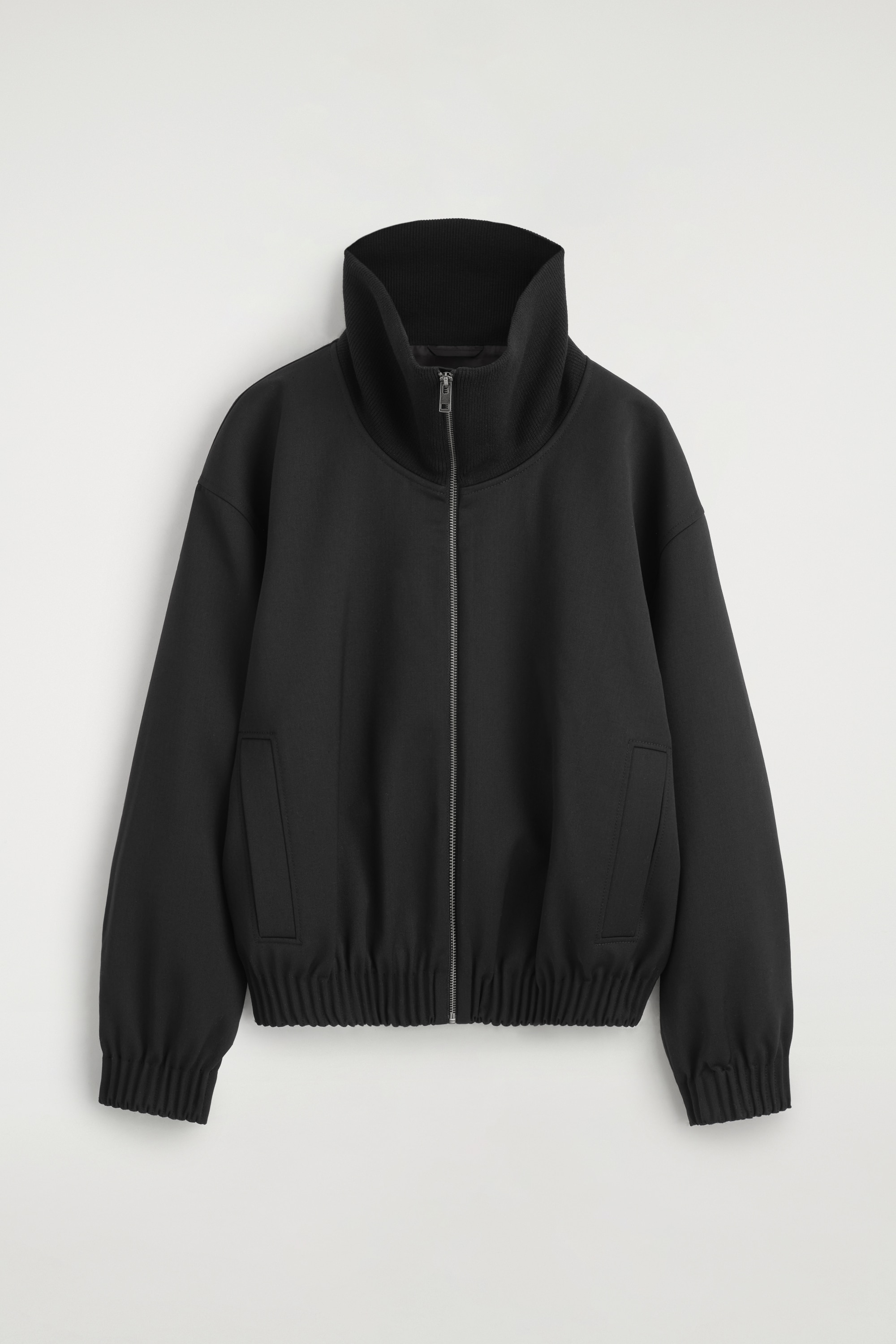 Den sorte jakke med funnel-neck bÃ¦res forfra og har en hÃ¸j krave, en sÃ¸lvfarvet lynlÃ¥s foran og en afslappet bomber-inspireret silhuet. Jakken har elastisk talje og manchetter, og modellen har hÃ¦nderne i sidelommerne.