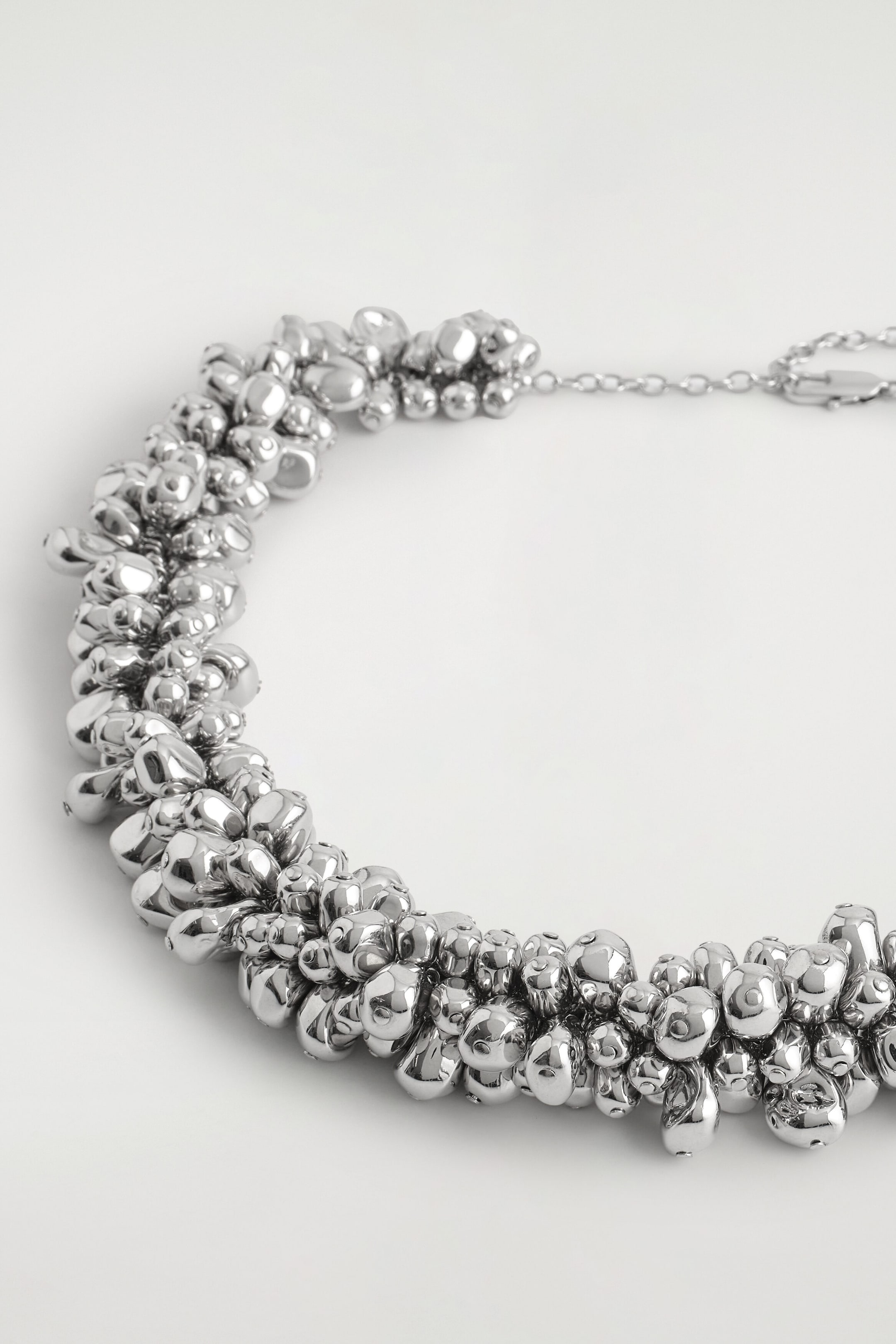 This silver-toned necklace features a thick, curved band made up of a dense cluster of polished, irregular metallic spheres. A delicate chain with a lobster-clasp fastening extends from the end of the clustered section. This silver-toned necklace features a thick, curved band made up of a dense cluster of polished, irregular metallic spheres. A delicate chain with a lobster-clasp fastening extends from the end of the clustered section.