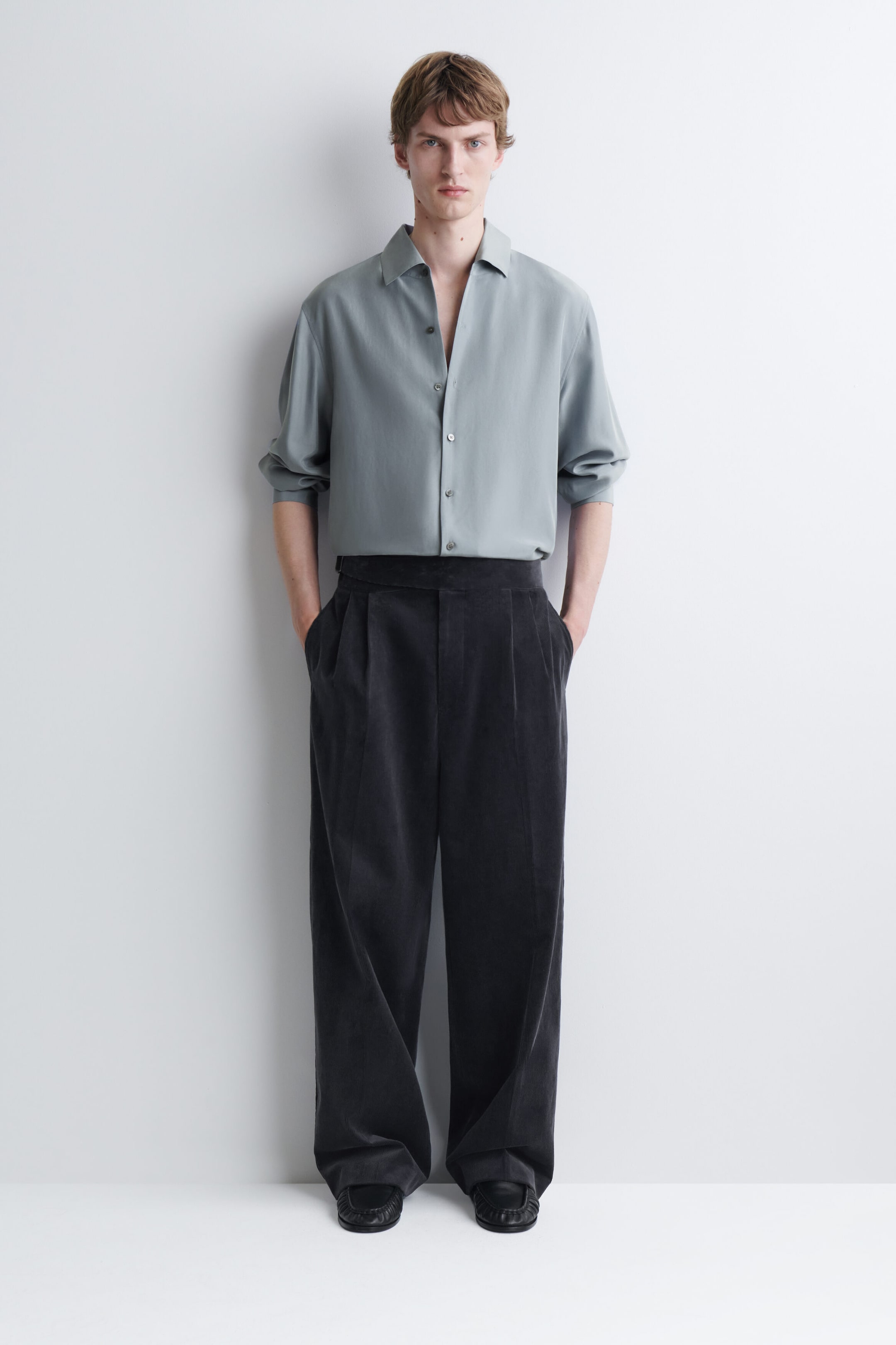 Belt-detail corduroy relaxed-fit wide-leg pants in a dark gray shade are being worn by the model, who is standing with hands resting in the side pockets. The high-waisted, pleated trousers are styled with a tucked-in, light gray button-up shirt and black shoes. Belt-detail corduroy relaxed-fit wide-leg pants in a dark gray shade are being worn by the model, who is standing with hands resting in the side pockets. The high-waisted, pleated trousers are styled with a tucked-in, light gray button-up shirt and black shoes.