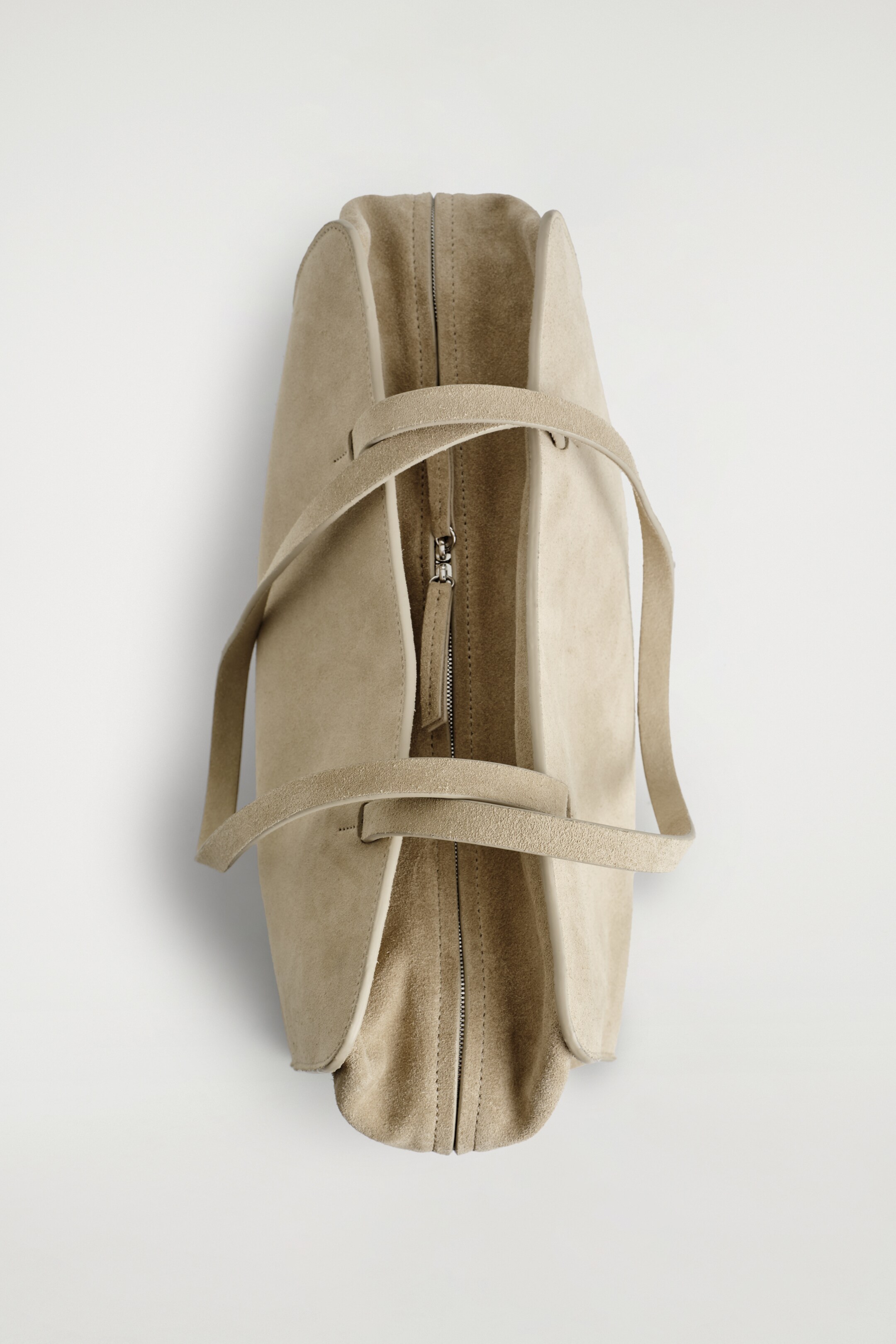 Zobrazit větší obrázek: Světle hnědá semišová bowlingová taška je zobrazena shora, má zapínání na zip s viditelným táhnutím, dvě ploché prodloužené popruhy a strukturovaný, zakulacený tvar.