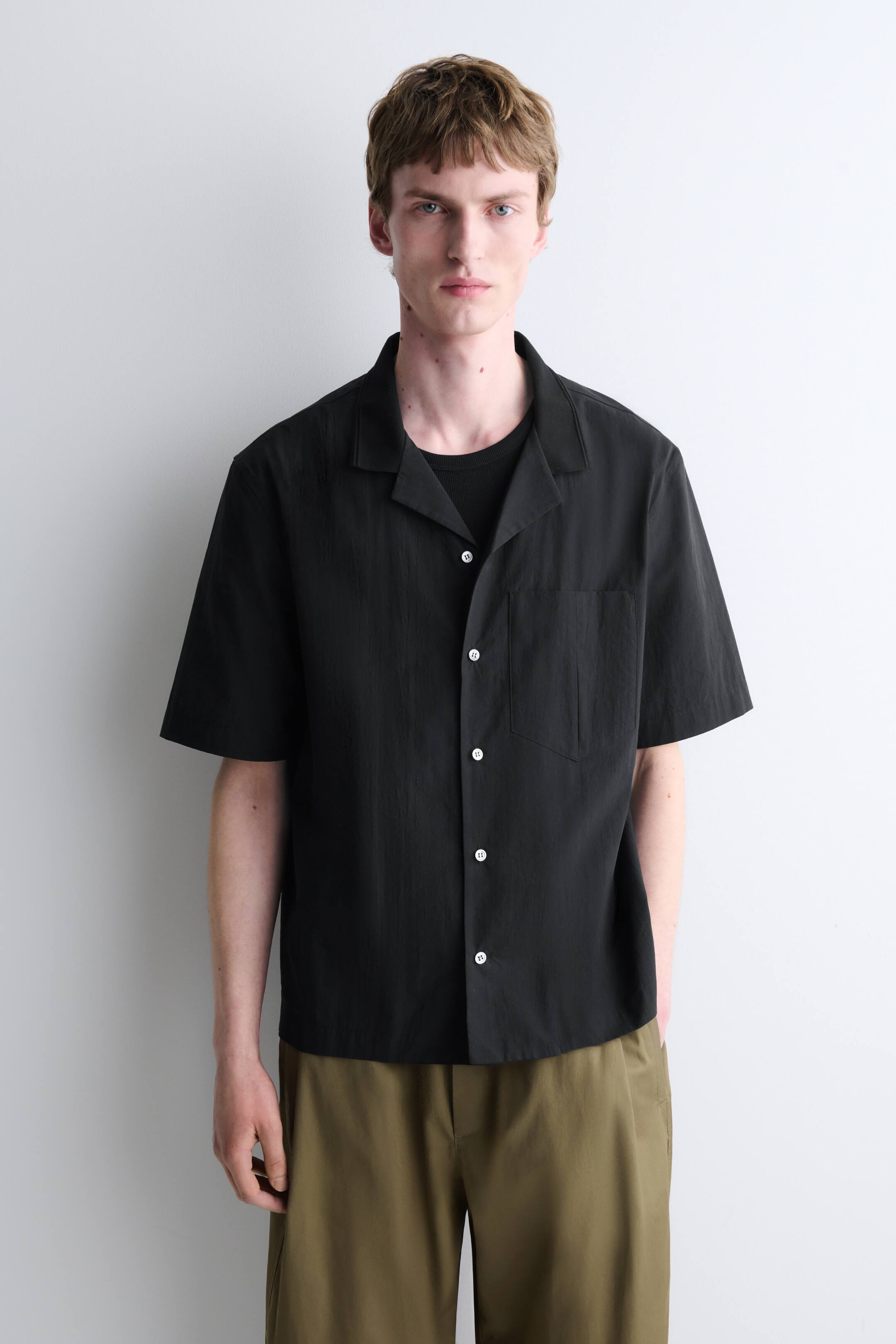 Größeres Bild anzeigen: Ein schwarzes Kurzarmhemd mit Camp-Kragen, weißem Knopfverschluss und einer Brusttasche wird vom Model getragen. Es ist offen über einem schwarzen T-Shirt gestylt.