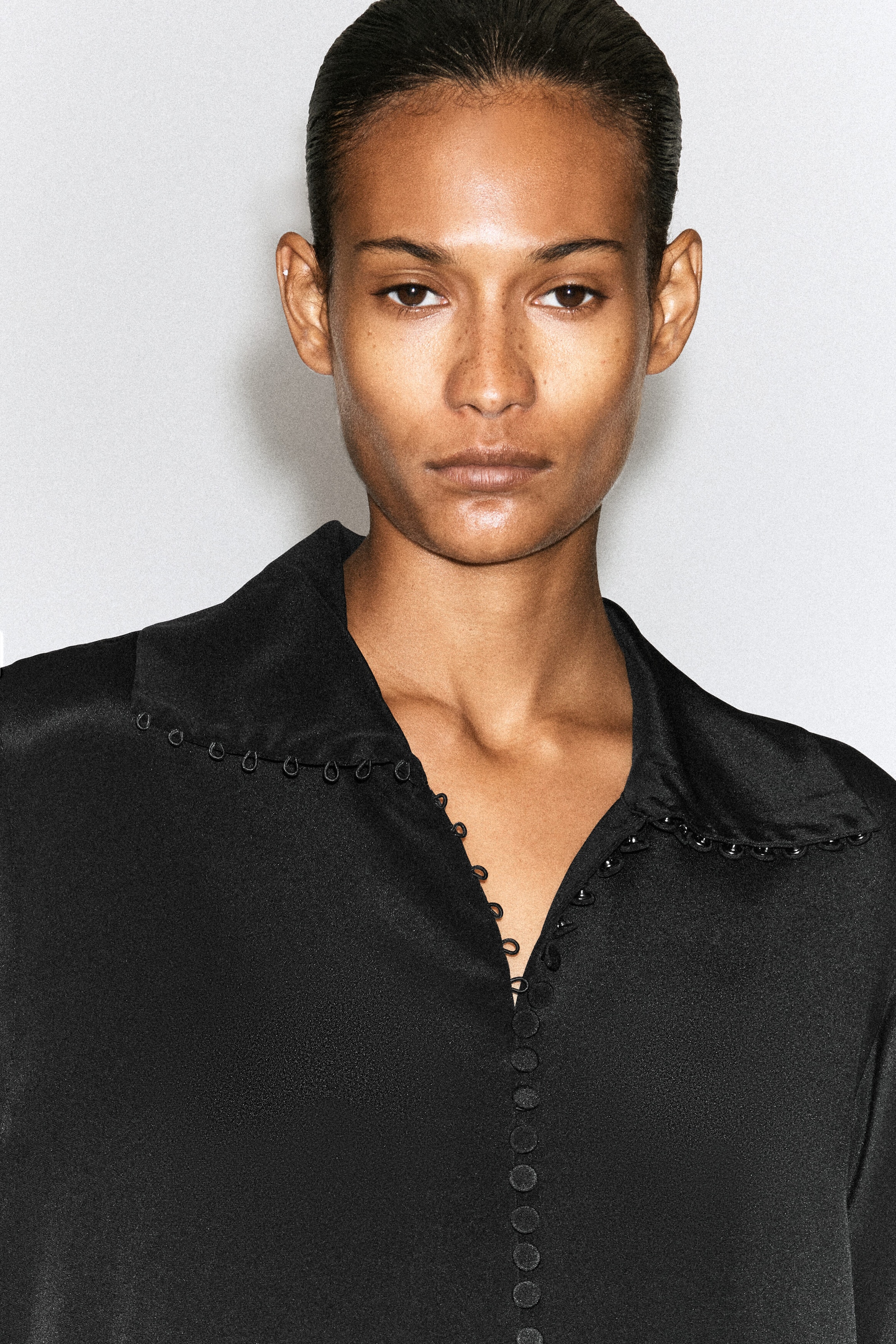 A black silk shirt featuring a soft collar and small, fabric-covered buttons with loop closures is being worn by the model. The shirt is shown partially unbuttoned at the front, highlighting the delicate button detailing along the collar's edge. A black silk shirt featuring a soft collar and small, fabric-covered buttons with loop closures is being worn by the model. The shirt is shown partially unbuttoned at the front, highlighting the delicate button detailing along the collar's edge.