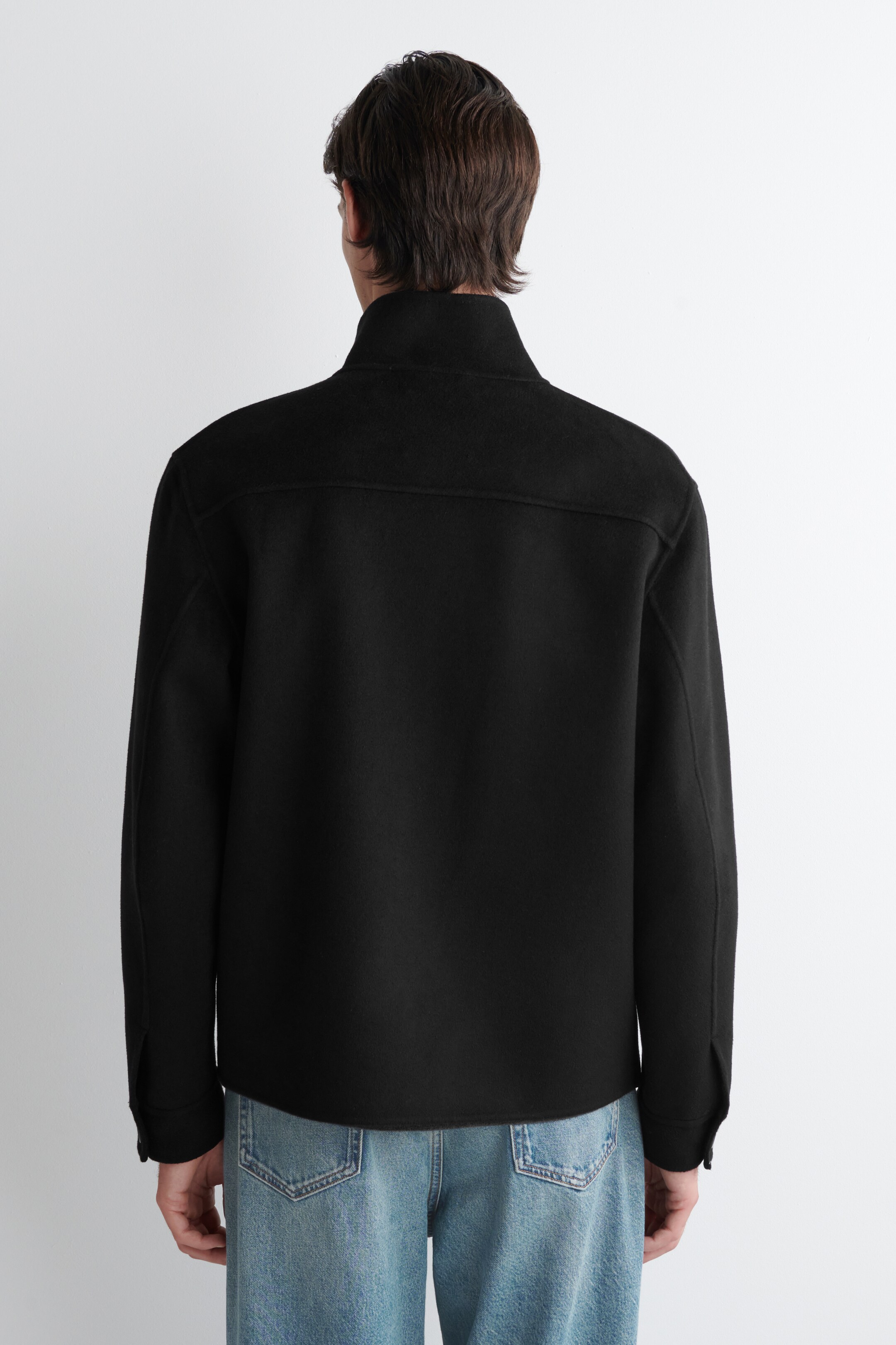 Προβολή μεγαλύτερης εικόνας: Ένα μαύρο σακάκι φαίνεται από πίσω, με όρθιο γιακά και μια οριζόντια ραφή τύπου ζυγού στο πάνω μέρος της πλάτης. Τα μακριά μανίκια του καταλήγουν σε κουμπωτές μανσέτες, και ένα ίσιο στρίφωμα αγγίζει ακριβώς πάνω από τη γραμμή της μέσης των μπλε τζιν.