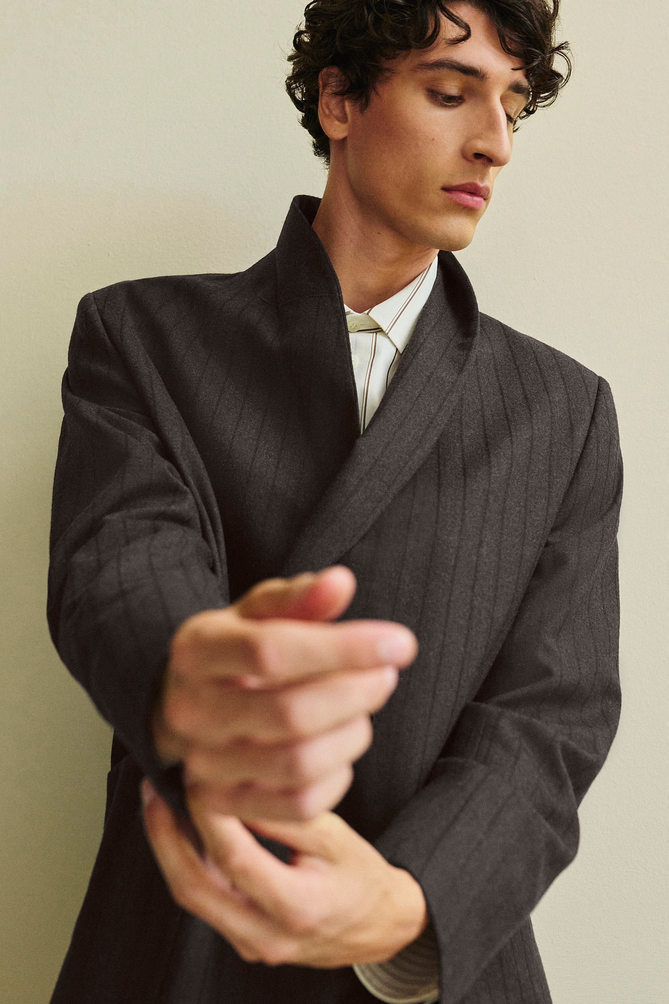 A dark grey double-breasted herringbone blazer with subtle vertical stripes is layered over a cream striped structured cotton-silk shirt. Shown from the front, the wearer is adjusting the blazer's cuff, highlighting the stand-up collar and the clean, structured drape of both garments. A dark grey double-breasted herringbone blazer with subtle vertical stripes is layered over a cream striped structured cotton-silk shirt. Shown from the front, the wearer is adjusting the blazer's cuff, highlighting the stand-up collar and the clean, structured drape of both garments.