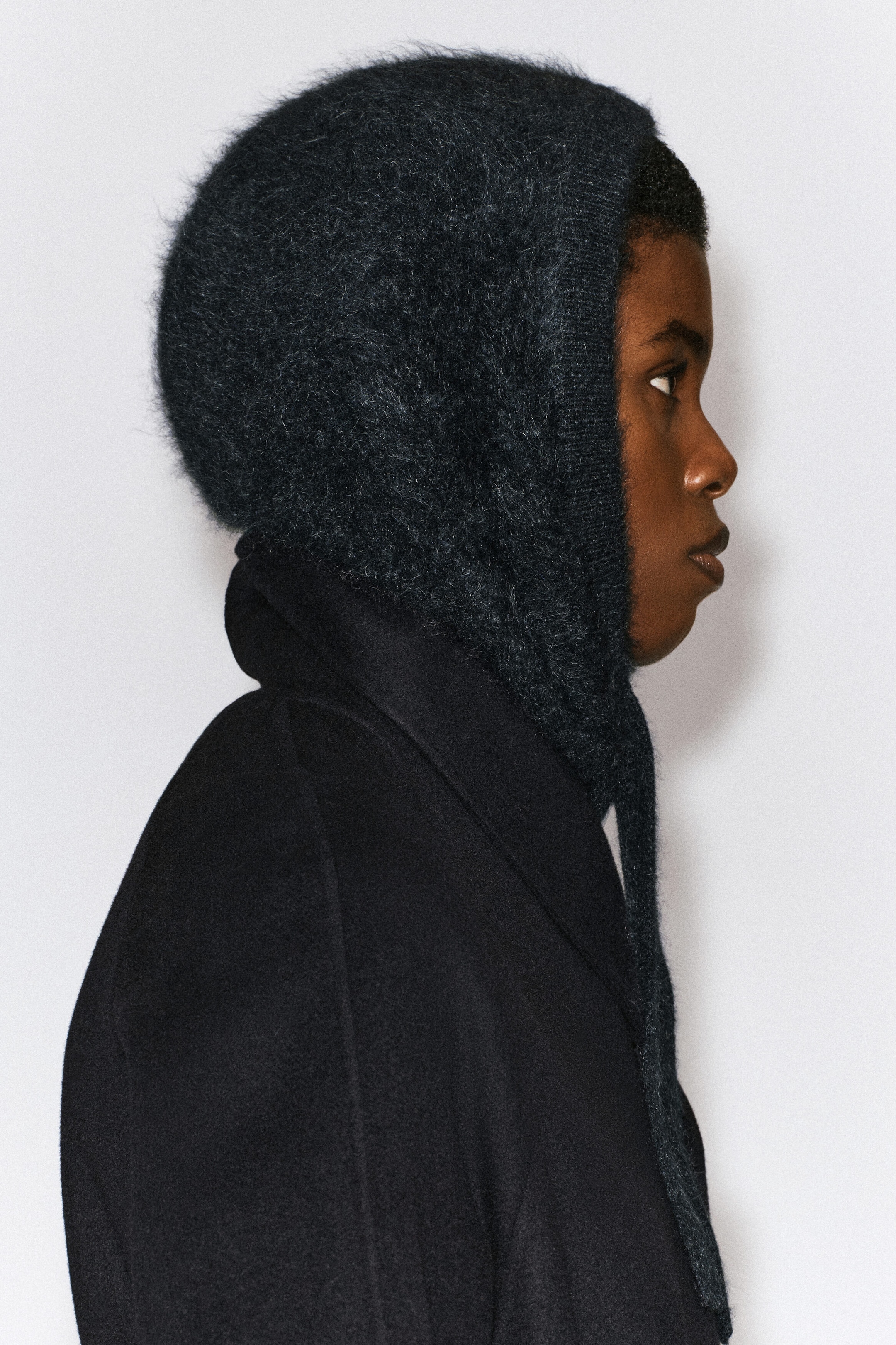 A dark gray brushed-knit hood is being worn by the model, shown in profile and layered over a black coat. The hood features a fuzzy texture, a ribbed border around the face, and long ties hanging down. A dark gray brushed-knit hood is being worn by the model, shown in profile and layered over a black coat. The hood features a fuzzy texture, a ribbed border around the face, and long ties hanging down.