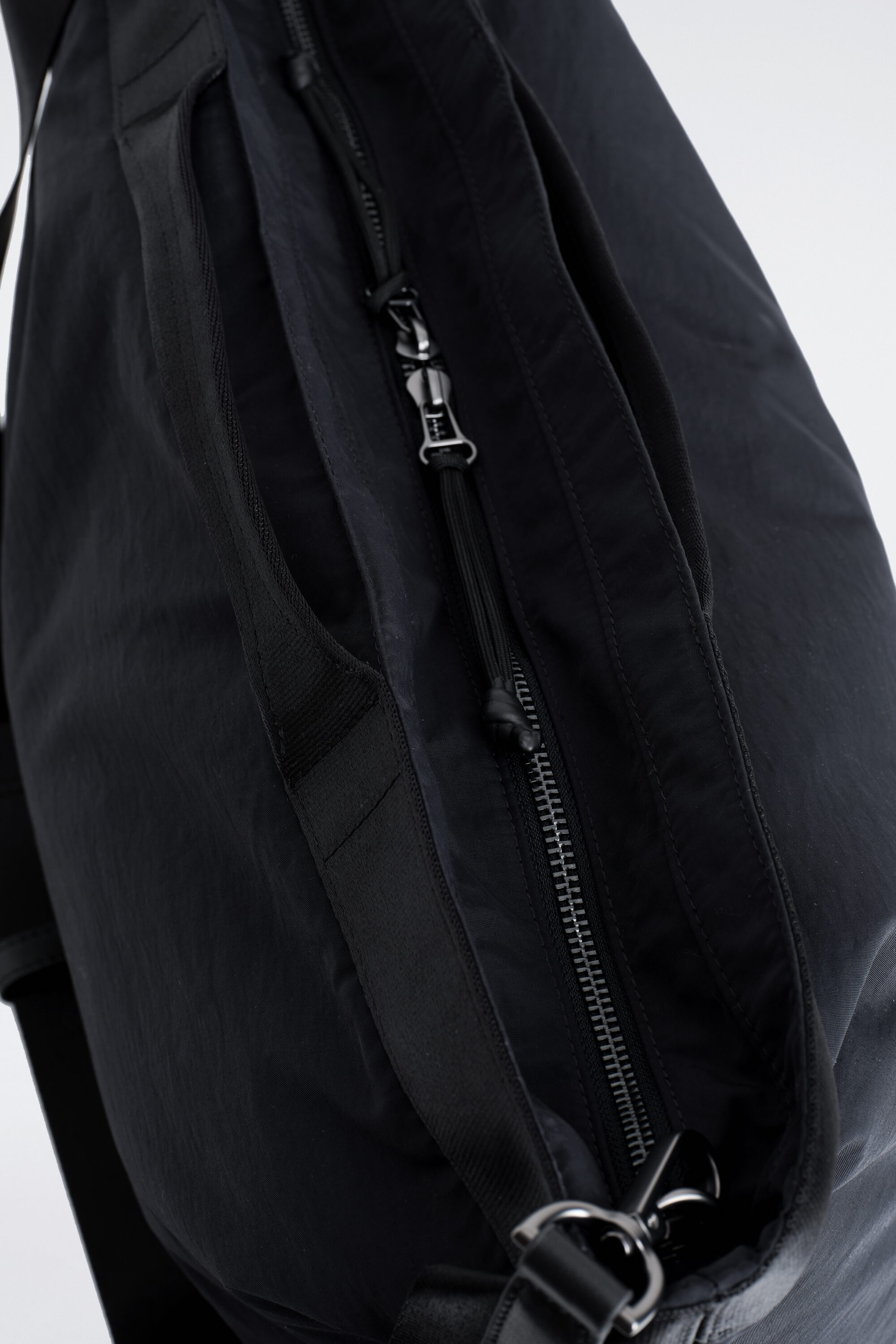 A black nylon bag is shown in a close-up view of its main zippered opening, featuring a metallic zipper pull and a long cord puller. A wide black webbing strap runs parallel to the zipper, leading to a metal ring attachment at the bottom.